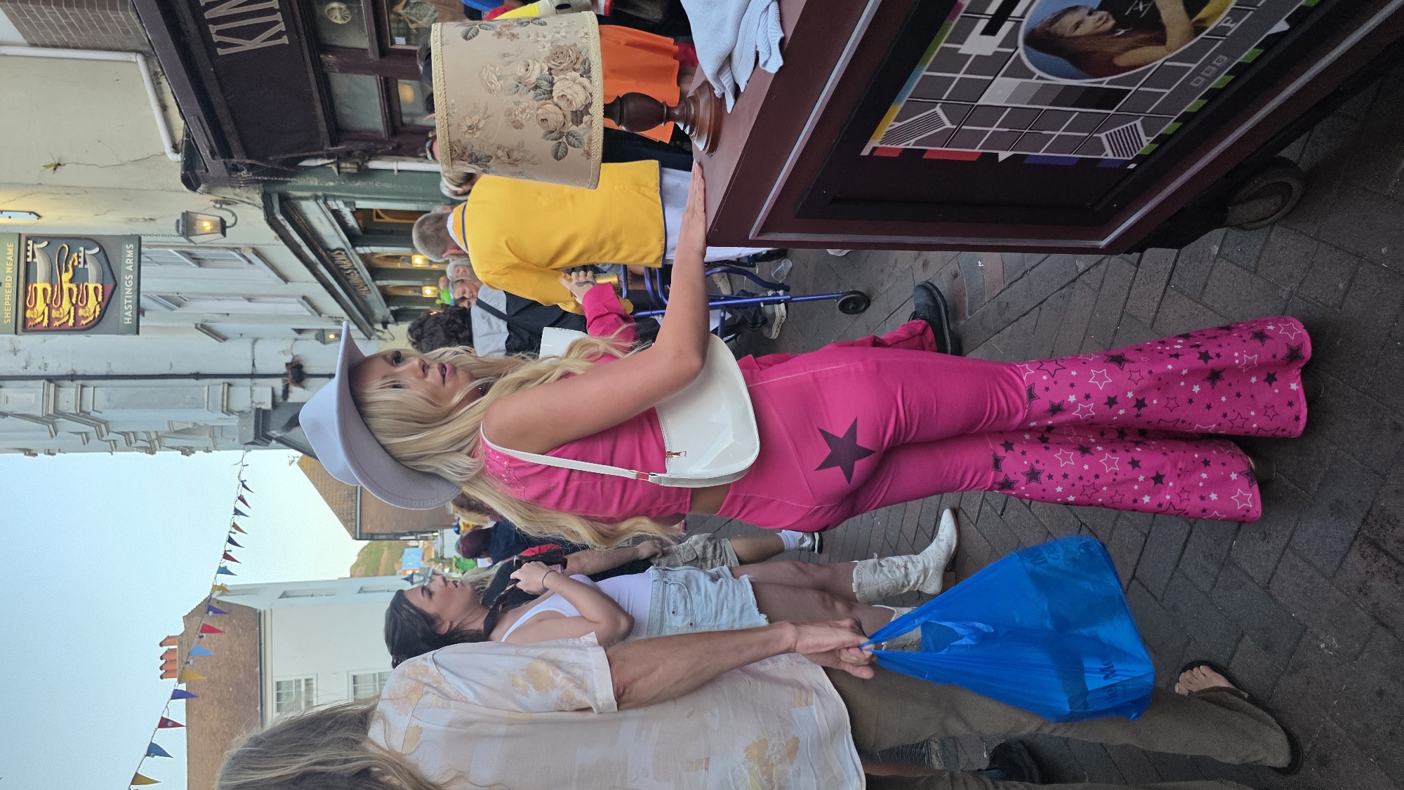 A young woman with blonde hair, wearing a white cowboy hat, a pink halter top, and pink bell-bottom pants adorned with black and white stars, walks down a street in St Leonards, United Kingdom. She carries a white shoulder bag. Behind her, a person in a yellow shirt pushes a walker. In the foreground, a man in a cream-colored shirt and tan pants walks to the left, holding a blue plastic bag. The street is paved with bricks, and buildings line both sides of the road, including a pub called "The Hastings Arms." Decorative bunting hangs across the street overhead. A vintage television set is displayed on a wooden stand to the right of the frame. The scene appears to be a street festival or event.