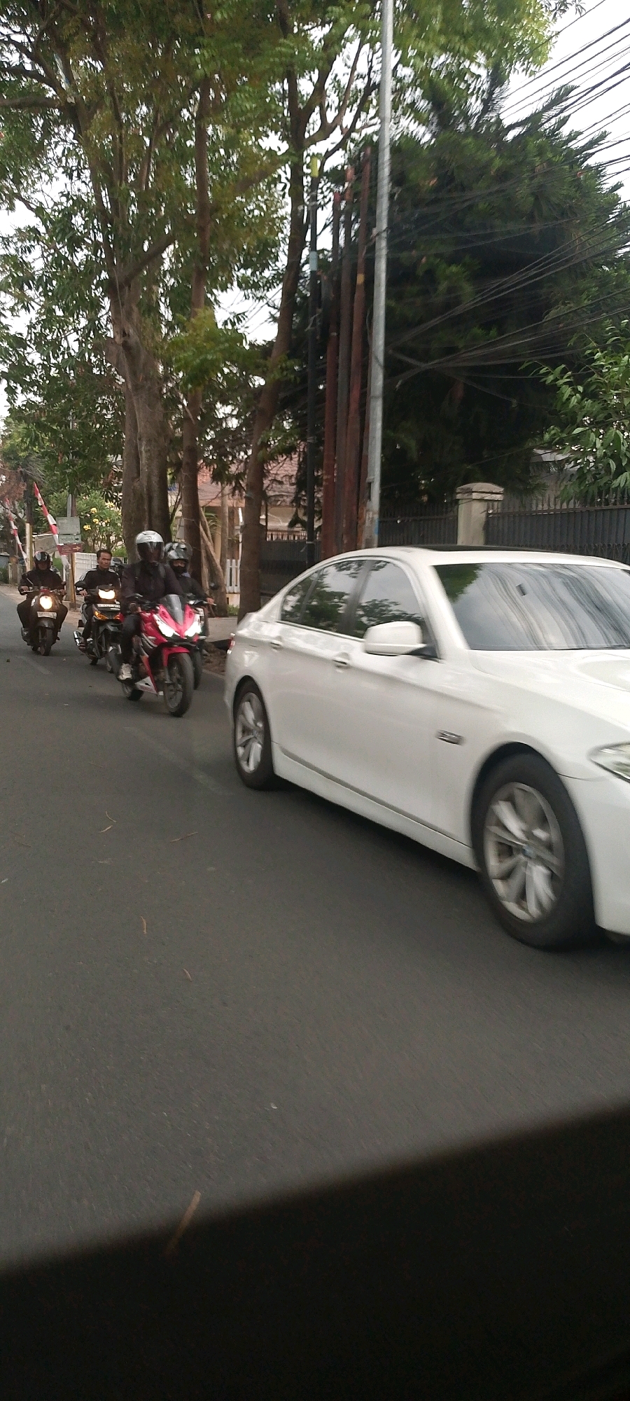 In Bandung City, Indonesia, a white sedan is captured in motion on an asphalt road, its wheels blurred by speed. The car is passing a line of motorcyclists traveling in the same direction. The first motorcyclist, closest to the camera, is on a dark motorcycle, followed by a motorcyclist on a red motorcycle. Further ahead, another motorcyclist is visible on a dark scooter. The background features lush green trees lining the street, with a power pole and wires also present. Buildings with tiled roofs are visible behind the trees. The scene depicts urban traffic flow.