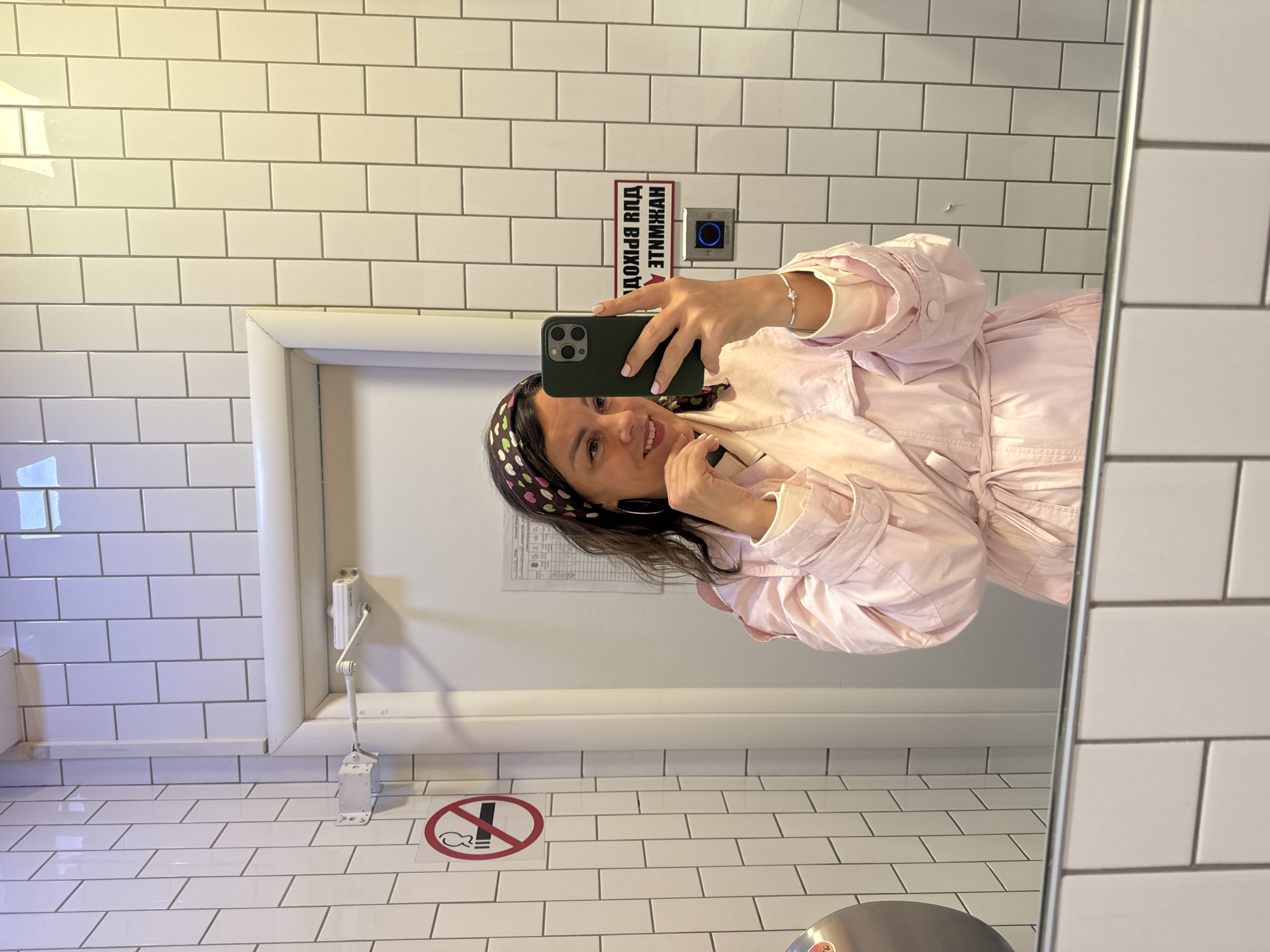 A woman is taking a selfie in a bathroom mirror. The bathroom has white subway tile walls. A sign in Cyrillic text is on the wall near an electronic touch button. The stall door is visible with a handicap assist bar and a sign prohibiting fishing. The bathroom is likely in Krasnoyarsk, Russia.