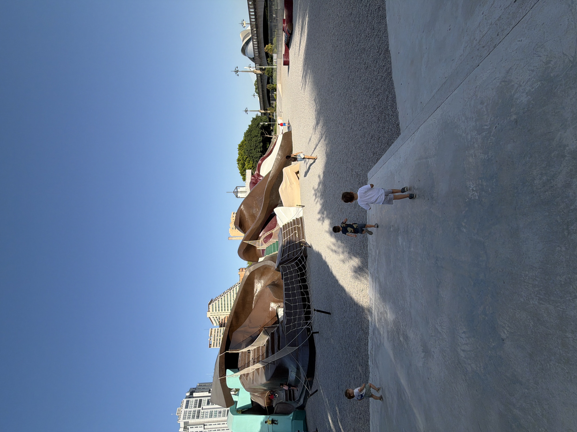The image depicts a sunny day at a modern playground in Valencia, Spain, with a clear blue sky. The playground features a large, abstract, brown and tan structure with slides and climbing nets, set against a backdrop of gravel and a smooth concrete area.
Several children are visible engaging in activities. One child in a white t-shirt and grey shorts walks on the concrete. Another child, also in a white t-shirt and grey shorts, stands near a red object. Two other children are seen playing on or near the playground structure. In the background, there are buildings, trees, and a large, modern stadium-like structure. The shadows suggest it is late afternoon. The overall mood is relaxed and recreational. No visible text is present.