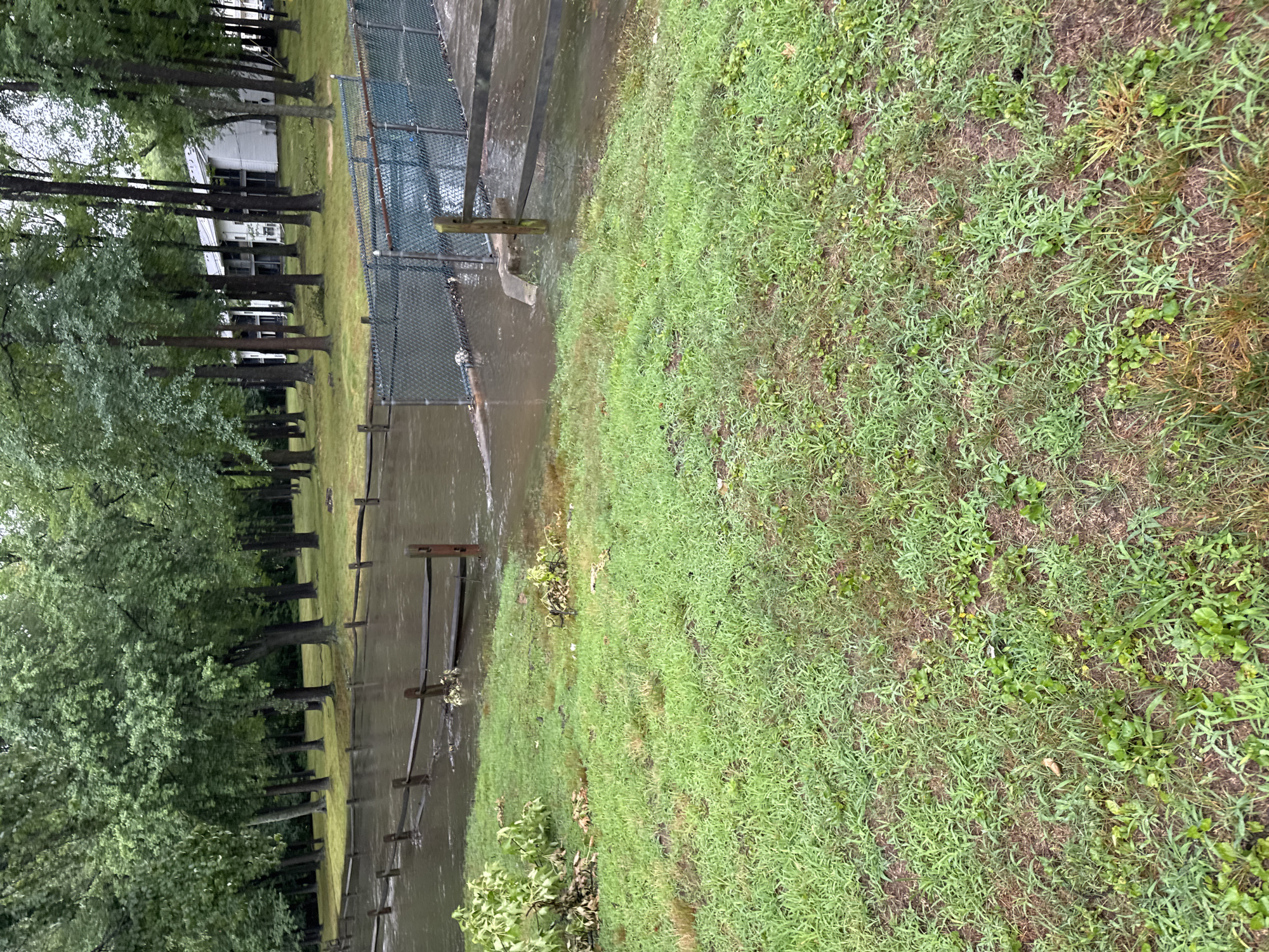 In Hamilton Township, United States of America, a flooded area is visible. A chain-link fence and a wooden fence run through the scene, separating a grassy area from the water. Trees line the background, and a white building with multiple stories is partially visible behind them. The ground is covered in grass, with some areas appearing muddy. Debris, including branches and leaves, is present in the flooded water and along the edge of the grass. The lighting suggests an overcast day.