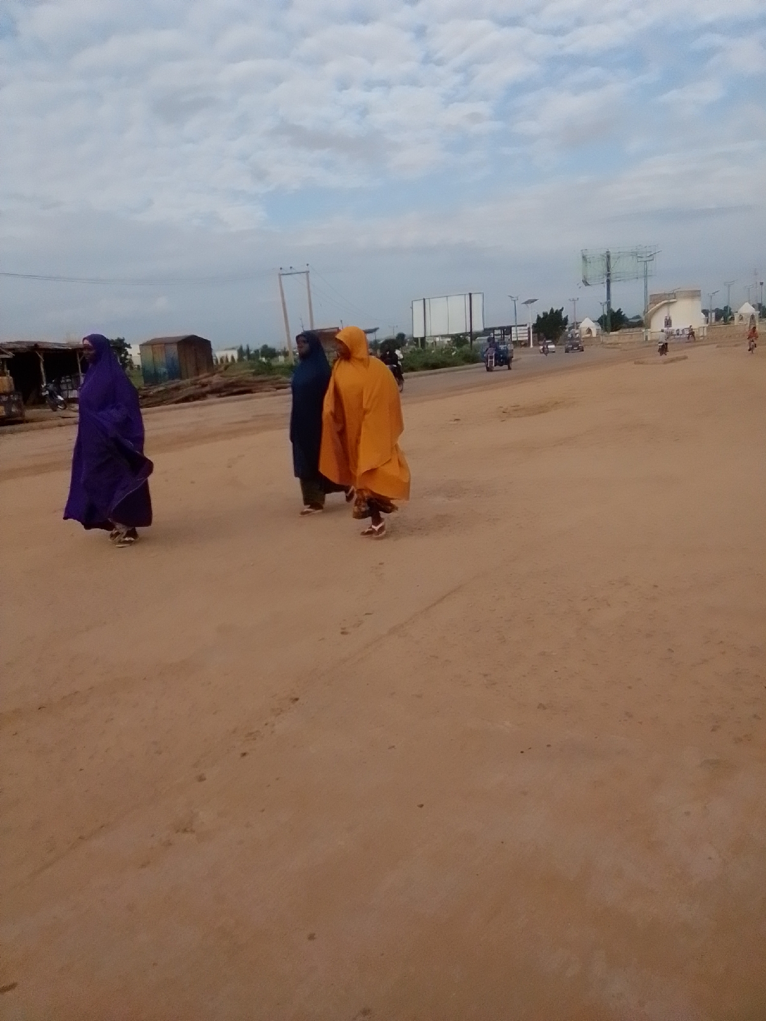 In Kwadon, Nigeria, three individuals are walking along a dirt road. The person on the left is wearing a purple hijab and abaya, the middle person a dark blue hijab and abaya, and the person on the right a bright orange hijab and abaya. The road is unpaved and dusty. In the background, there are structures that appear to be market stalls or small shops, along with a few trees and utility poles. Further in the distance, there are buildings, billboards, and vehicles on a paved road, suggesting the scene is on the outskirts of a more developed area. The sky is cloudy.