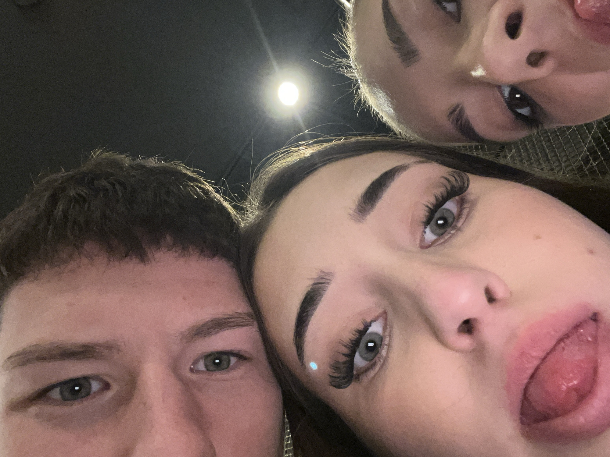 Three people pose for a selfie. A man with short brown hair and green eyes is on the left. Two women with long, dark hair and green eyes are to the right and above him. One woman has her tongue out, and the other has a small white mark on her nose. A light is visible in the background. The location is not discernible from the image.