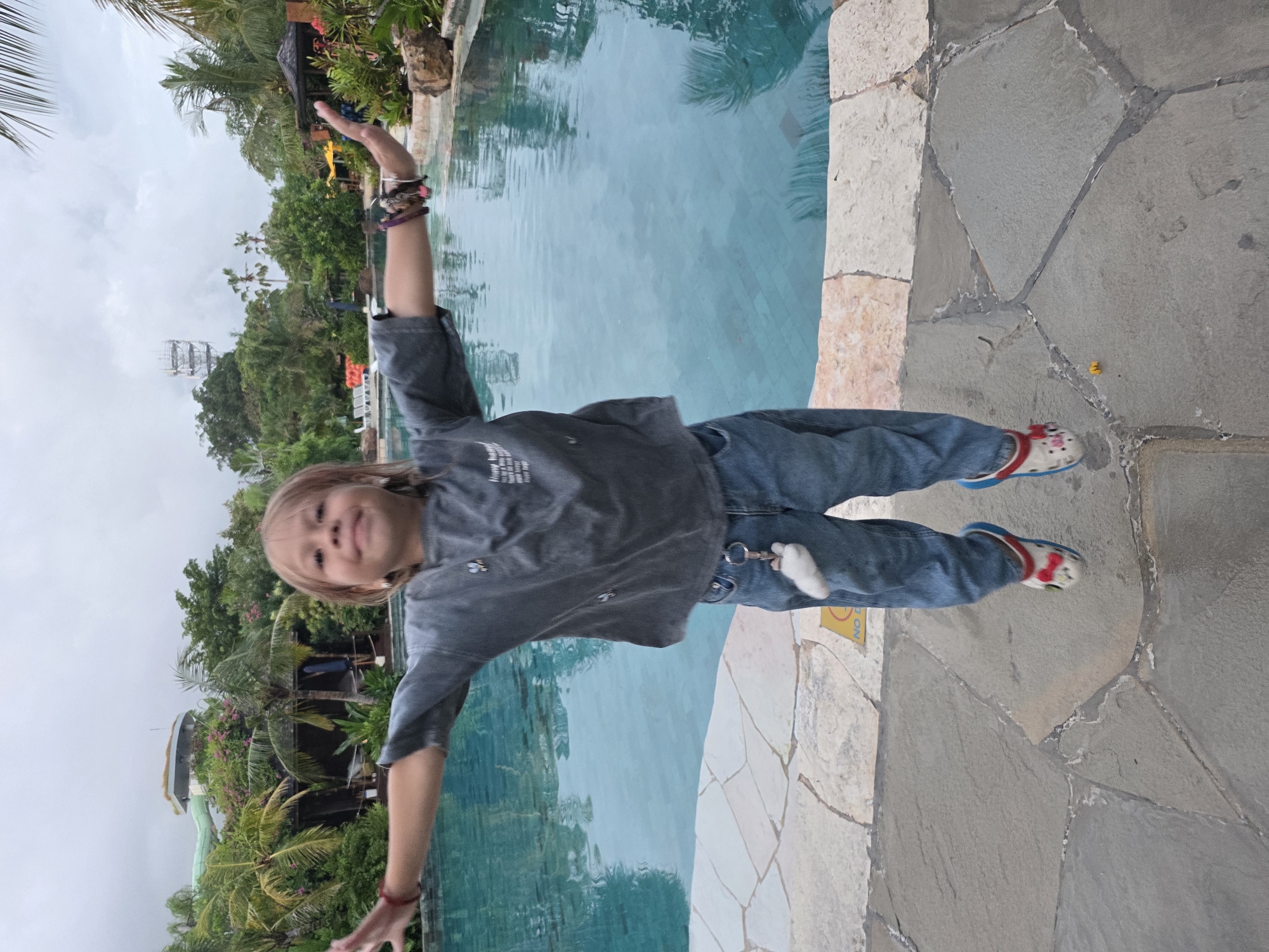 A young child stands with arms outstretched next to a swimming pool, at a location in Kuta, Indonesia. The child wears jeans, a gray shirt, and patterned Crocs. The pool has light blue water and a natural stone edge. Lush tropical vegetation is visible in the background, including palm trees and other greenery. A tower-like structure is noticeable in the background behind the trees. The ground surrounding the pool is paved with irregular stone slabs. A fluffy, cloud-like object hangs on a carabiner attached to the child's jeans.
