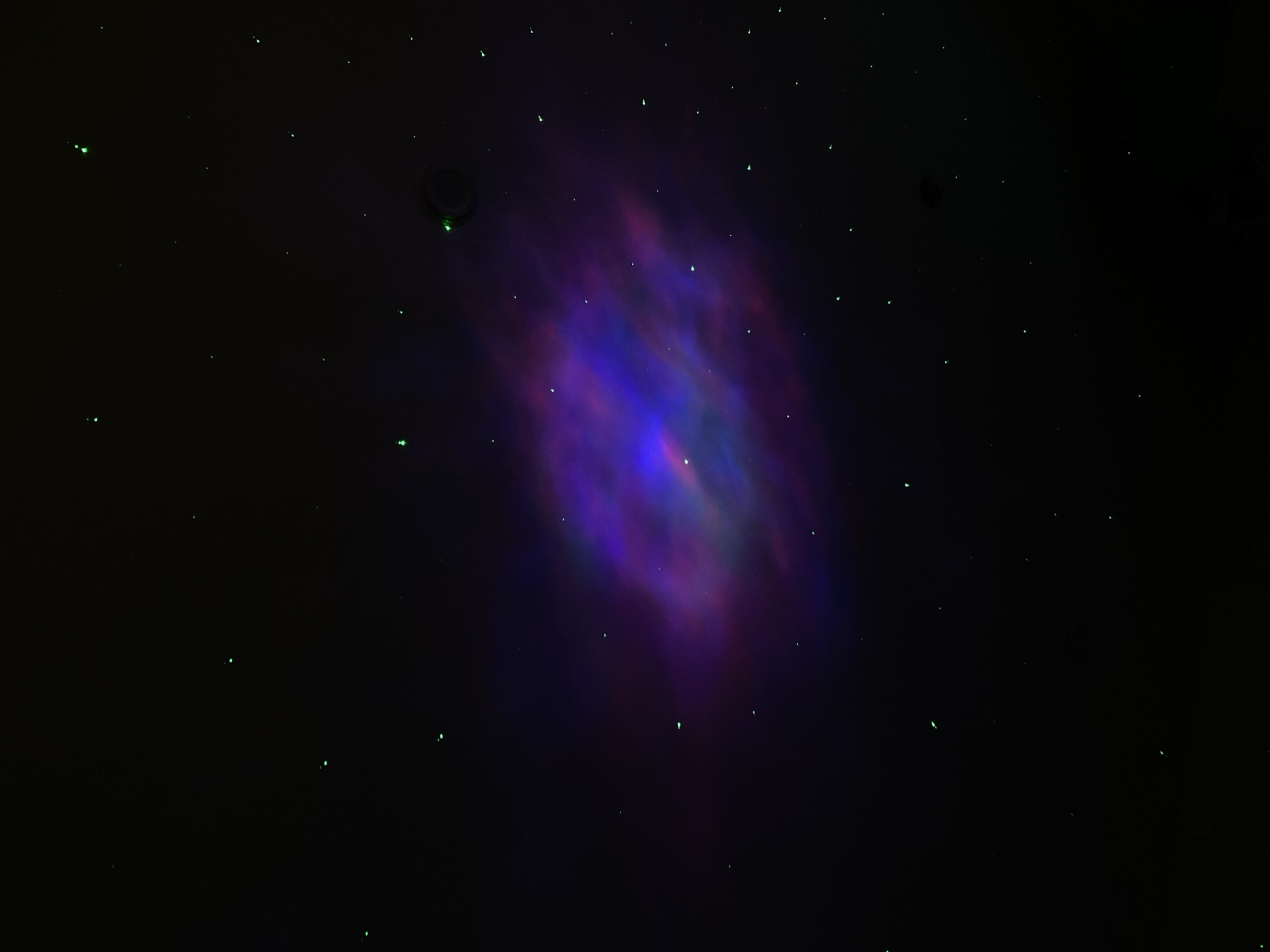 The image shows a dark night sky with a prominent nebula-like formation of blue, purple, and reddish hues. Scattered across the black background are numerous small, bright green specks, resembling stars. Two dark, circular shapes are visible in the upper part of the image. There are no people or specific objects identifiable that would indicate the location being Votkinsk, Russia.