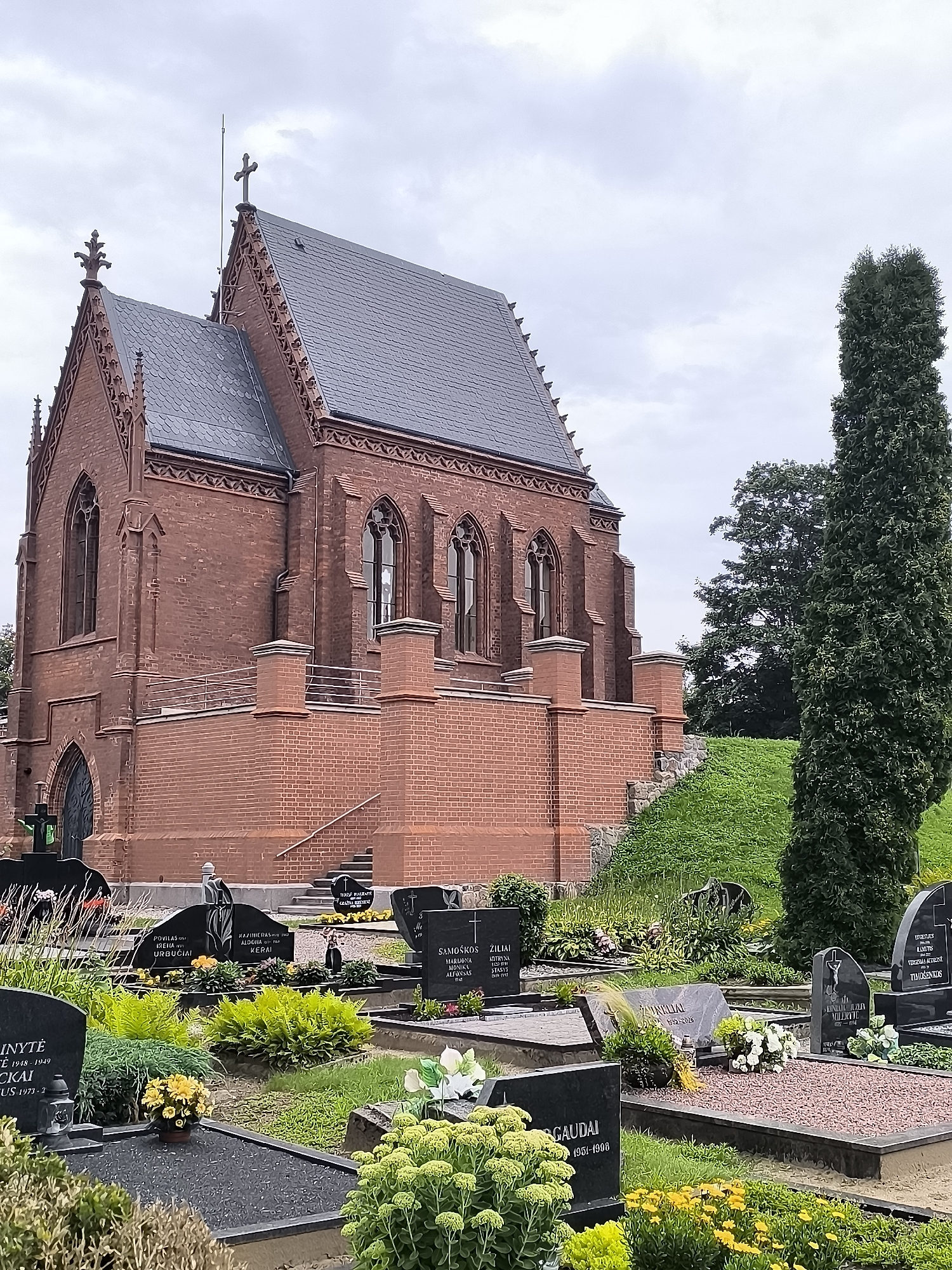 This is a photograph of a cemetery in Kretinga, Lithuania, featuring a prominent red brick mausoleum with Gothic architectural elements. The mausoleum has a steep, grey-tiled roof, pointed arched windows, and decorative finials. A tall, dark green evergreen tree stands to the right of the mausoleum. In the foreground, numerous gravestones of varying sizes and materials, many with inscriptions and floral arrangements, are arranged in rows. The ground is covered with grass and various low-lying plants and flowers. The sky is overcast with grey clouds. The overall impression is one of solemnity and remembrance.