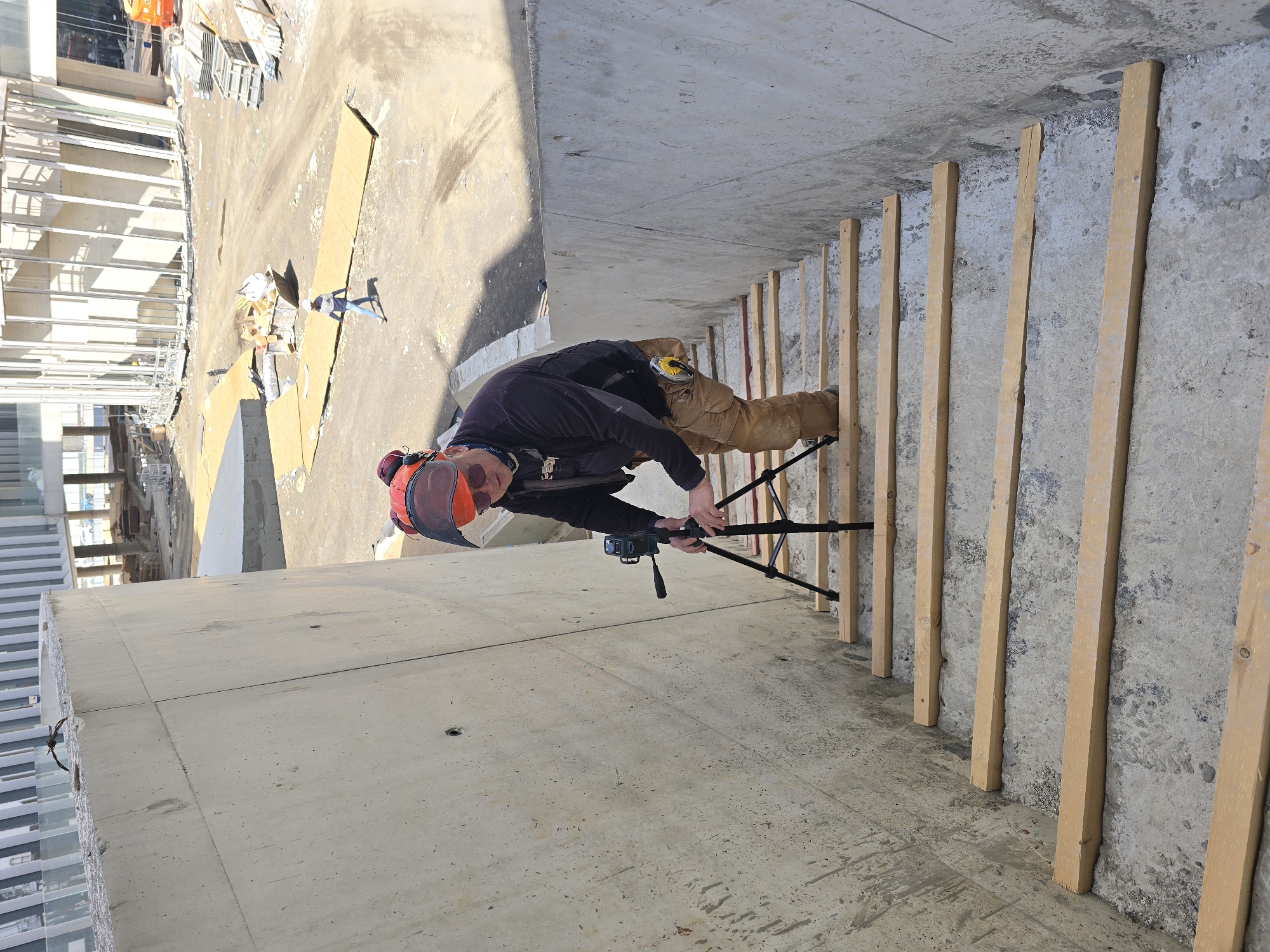 A person with an orange safety helmet and a dark jacket stands on a concrete surface, possibly an unfinished balcony of a building under construction, with wooden planks lining the edge. The person is operating a black tripod with a camera mounted on top. In the background, the urban environment includes buildings and construction materials, suggesting a city location, potentially in the Unknown. Another person is visible in the distance with a blue shirt.
