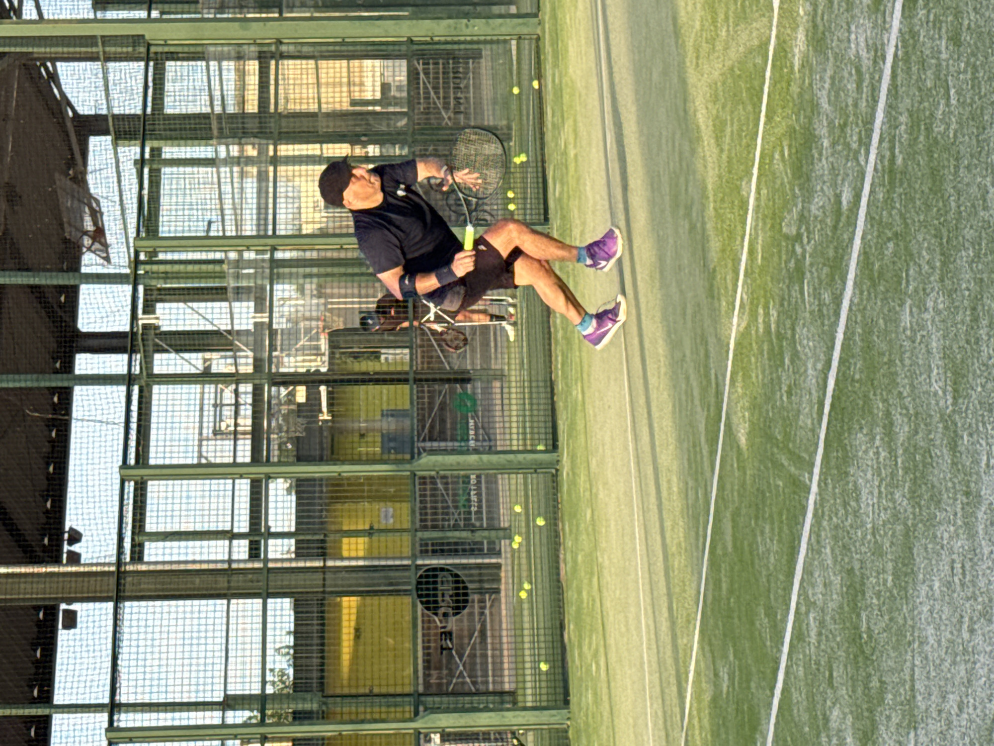 A man wearing a black t-shirt, dark shorts, a black cap, purple athletic shoes, and a black arm brace on his left arm is playing padel on an outdoor court. He is in mid-action, bending his knees and holding a padel racket in his right hand, poised to strike a yellow ball. Several yellow padel balls are scattered on the green court surface behind him. The court is enclosed by green metal fencing with netting. In the background, another person is visible, also holding a racket. The court is situated within a larger structure with a metal roof and vertical supports, suggesting it's part of a sports complex in Vidreres, Spain. The lighting indicates it is daytime, with sunlight casting shadows on the court.