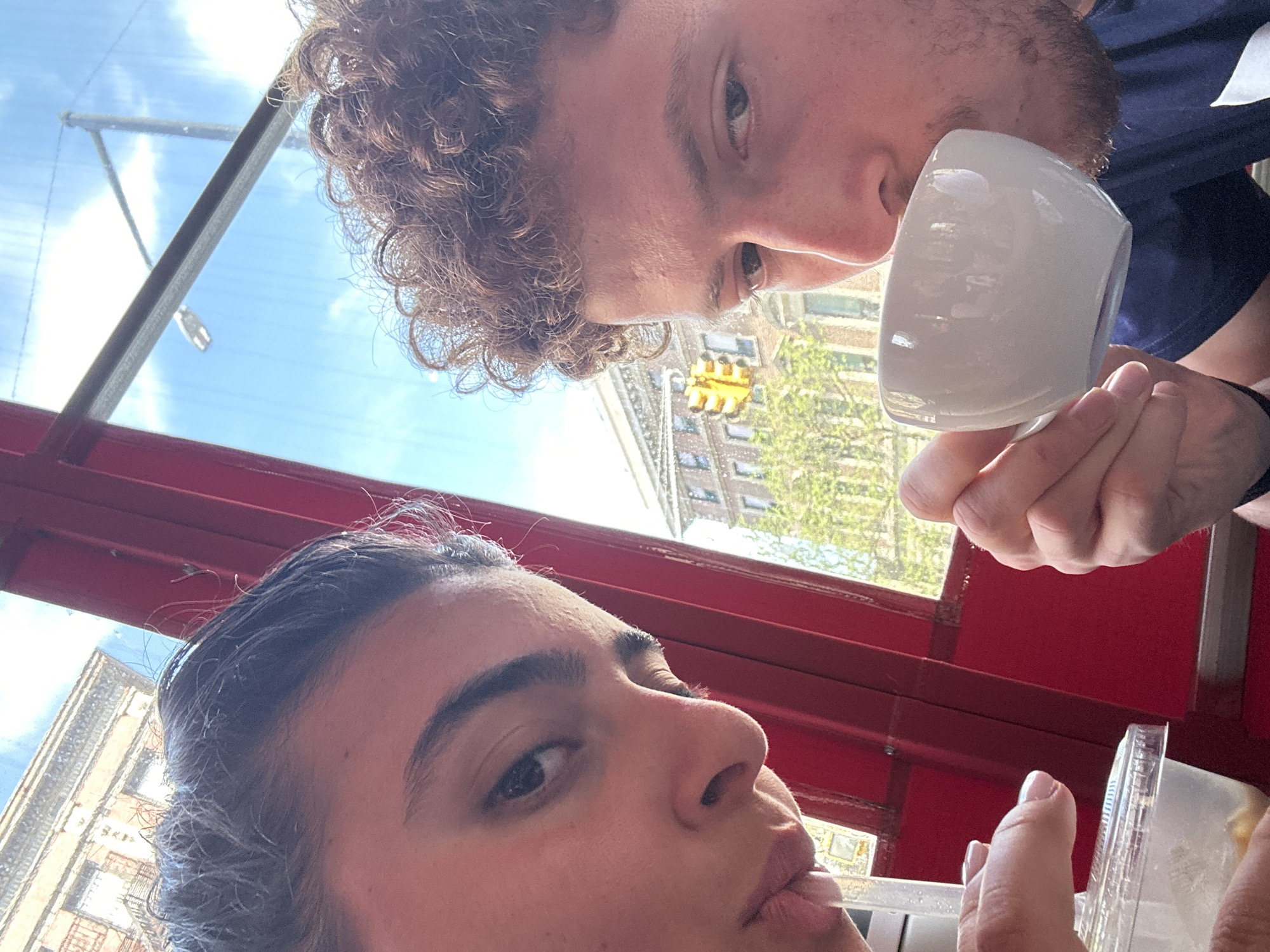 This close-up, inverted selfie-style photograph features two individuals positioned in the foreground. The person at the top of the frame has light brown, curly hair and is captured mid-sip from a white, handleless mug, with their eyes directed towards the camera. Below them, another person with dark hair is partially visible, holding a clear plastic container, possibly a drink, and looking towards the lens.
The setting appears to be indoors, looking out through a large window framed by distinctive red sills. The outdoor scene reveals a bright, clear daytime sky with hints of clouds, suggesting sunny weather. An urban landscape is visible through the window, featuring multi-story buildings with windows, vibrant green trees, a yellow traffic light suspended over an intersection, and what appear to be utility poles or streetlights. The overall impression is a casual moment, possibly at a cafe or restaurant, overlooking a city street. No discernible text is visible in the image. Given the context, the scene is in an urban area of New York, USA.