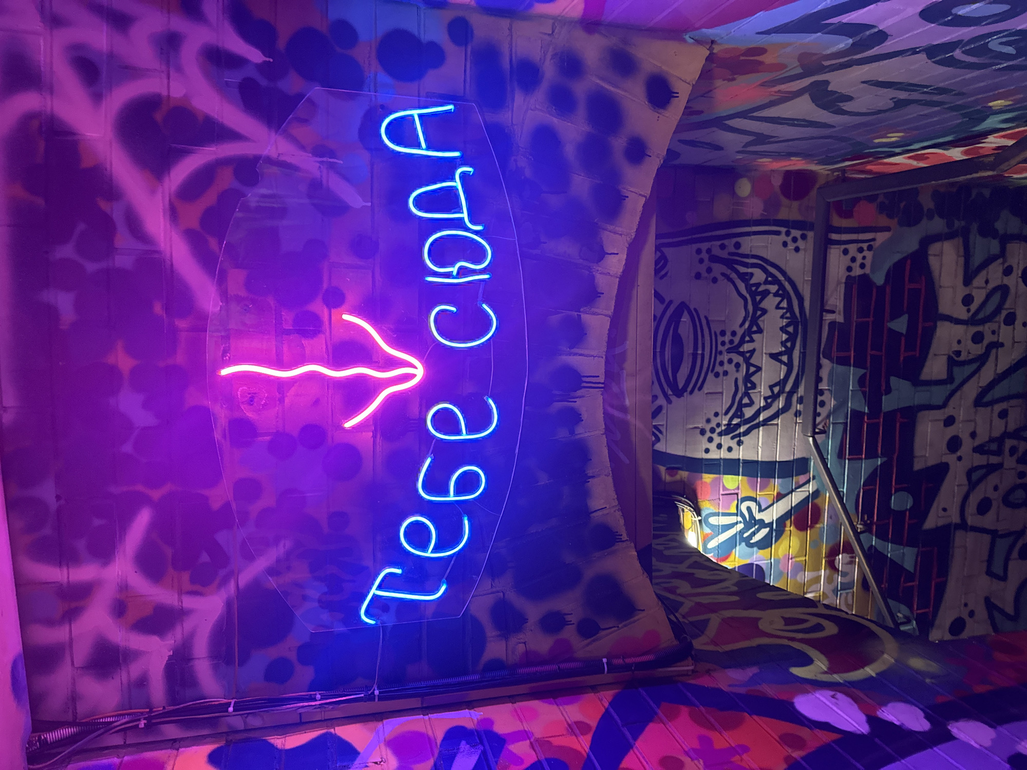 The image shows a neon sign in Russian that reads "Тебе сюда" (which translates to "You are here" or "This way for you") and an arrow pointing downwards. The sign is illuminated in bright blue neon and is mounted on a brick wall covered in graffiti. The environment appears to be an indoor space, possibly a stairway or tunnel, with more graffiti visible on the walls and ceiling. The lighting is dim, with purple and pink hues from the neon signs and possibly other light sources. The overall aesthetic is urban and edgy, suggestive of a nightlife venue or a creative space. The image is taken from a low angle, looking up towards the neon sign. There are no people or animals visible in the image. The location in Krasnoyarsk, Russia is not identifiable from the image itself.