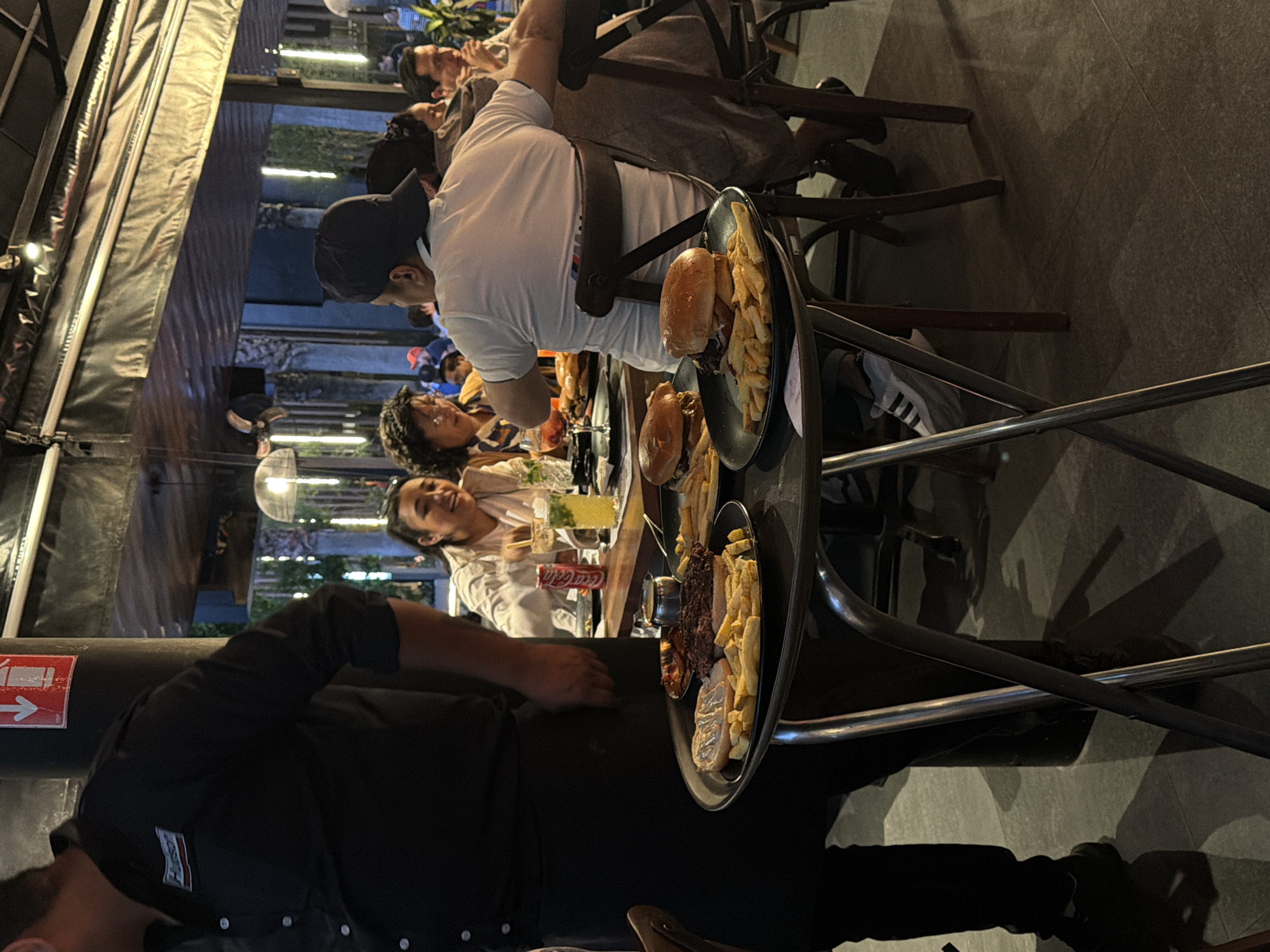The image shows an outdoor dining area, possibly in San Bernardino Tlaxcalancingo, Mexico. A waiter in a black uniform is carrying a multi-tiered tray of food, likely burgers and fries, towards a table occupied by a group of people. The diners are engaged in conversation and eating. The scene is set outdoors under a tent-like structure. The lighting appears to be artificial. The presence of drinks, including what looks like a Coca-Cola bottle and a cocktail, suggests a social gathering.