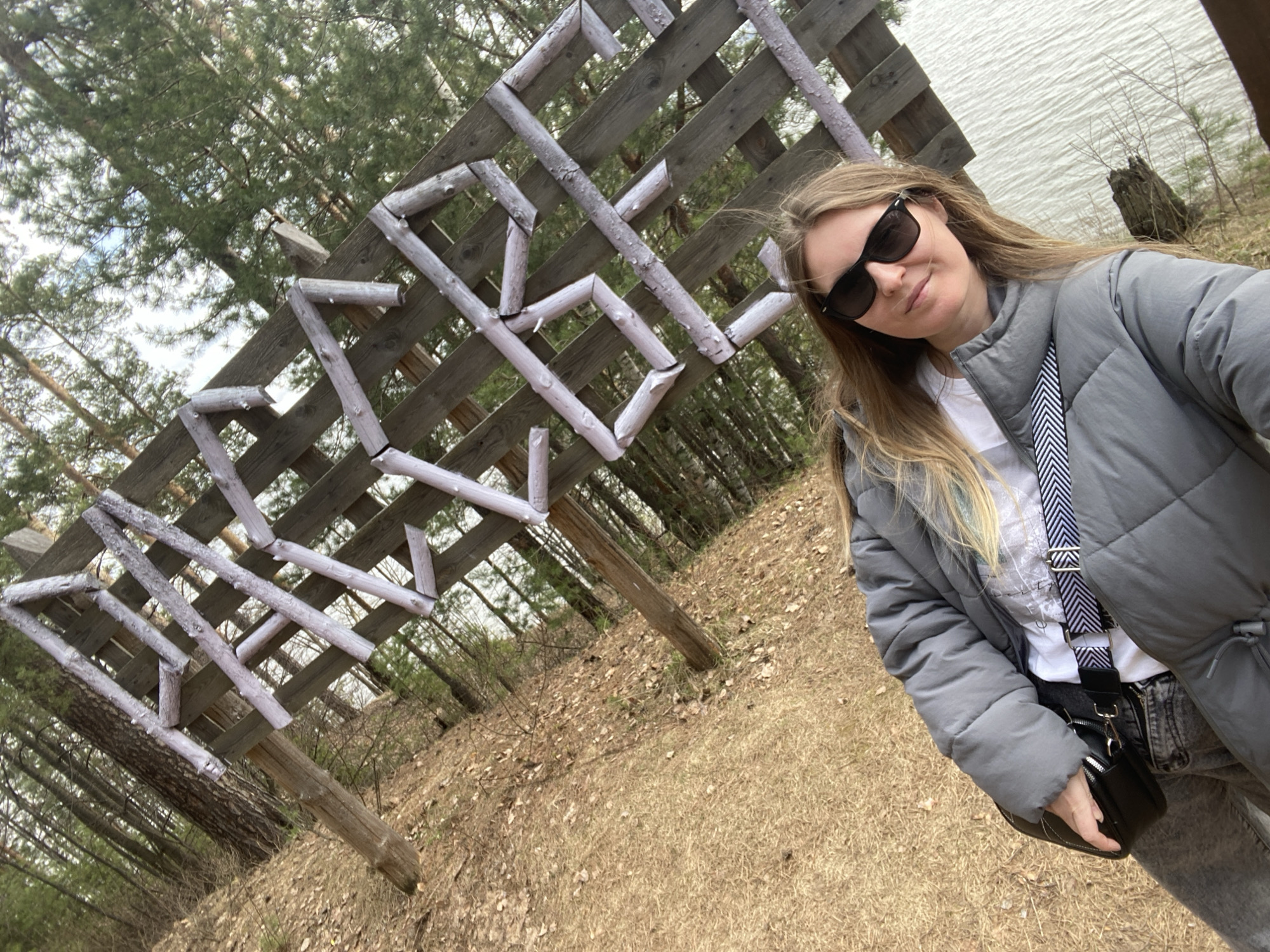 The image shows a woman taking a selfie outdoors, likely in a natural setting with a lake in the background. The woman is wearing a gray puffer jacket, sunglasses, and a crossbody bag with a black and white strap. Behind her is a wooden structure with a lattice pattern, possibly a sign or artwork. The ground is covered with dry grass and leaves, and trees are visible in the distance. The location is in Noviy, Russia.