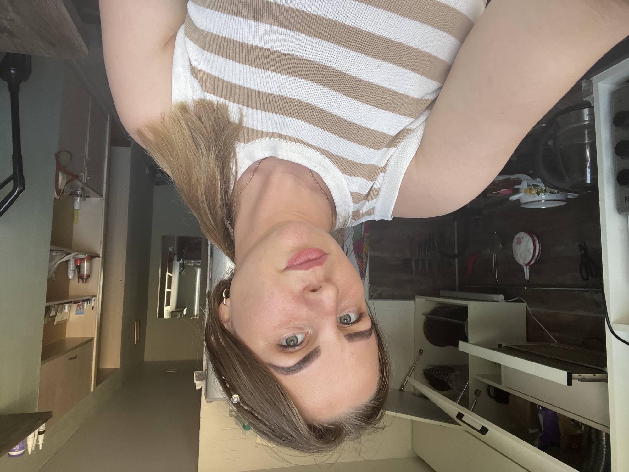 A woman with light skin, light brown hair, and green eyes is positioned centrally in the image, wearing a horizontally striped white and tan top. Her face is visible and slightly tilted. The background contains a kitchen area with white cabinets and various kitchen appliances. In the background, the hallway leads to cabinets containing toiletry items. There's no definitive visual element indicating that the location is Izhevsk, Russia.