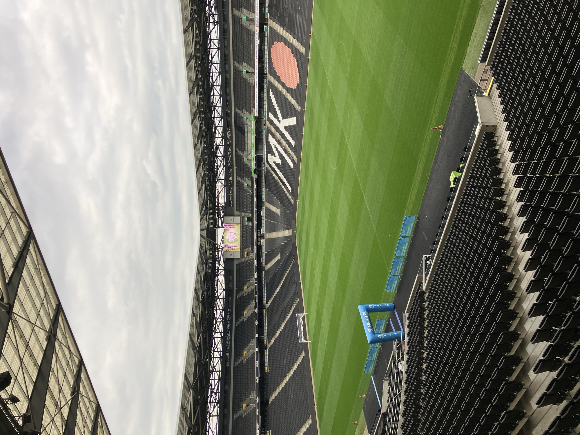The image shows a partial view of Stadium MK in Milton Keynes, United Kingdom. The stadium is largely empty, save for a few individuals near the field. The green field is striped with slightly darker lines. Blue arches labeled "MK Finish 5K" are placed near the edge of the field. A goal post is visible near the far end of the field. Black seats occupy most of the visible stands, with some white and red decorative elements on one side. The sky is overcast. Structural elements of the stadium roof are visible on both sides of the frame.