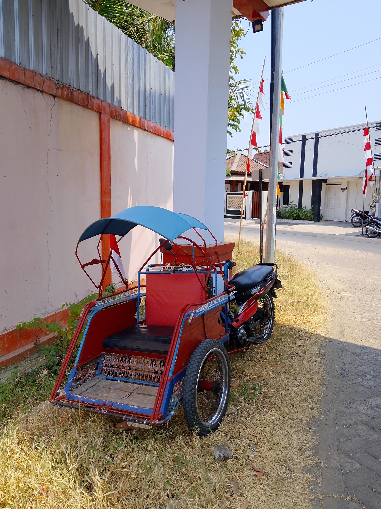 An abandoned red and blue motorized rickshaw is parked on dry grass beside a paved road in Biting, Indonesia. The rickshaw has a blue canopy, a black seat, and two wheels at the rear. A motorcycle is parked behind the rickshaw. Several Indonesian flags are displayed on poles in the background, near a white building with black vertical stripes. The area appears to be a residential street.