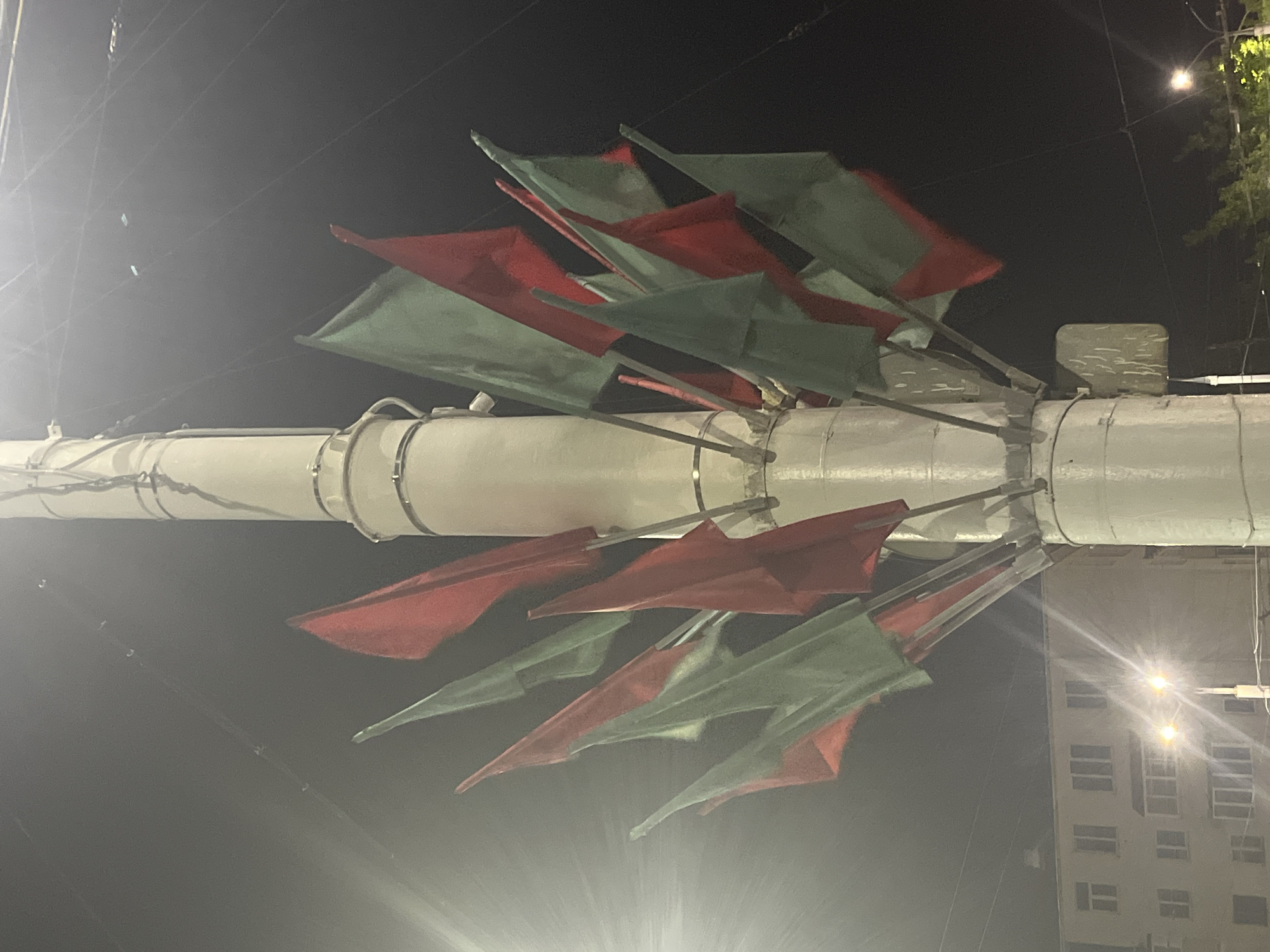 The image shows a lamppost adorned with numerous red and green flags at night. The lamppost is white and cylindrical, with several circular metal bands wrapped around its base. A cluster of flags, appearing to be made of fabric, are attached to the lamppost at various angles, some pointing upwards and others downwards. The flags alternate in color between red and green. The background is dark, suggesting it is nighttime. In the lower left portion of the image, a light-colored building with multiple windows is visible. Two streetlights are also present, casting a bright glow and creating lens flare effects. The scene is likely an outdoor urban environment. The specific location in Vitsebsk, Belarus, is not identifiable from the visual information alone.