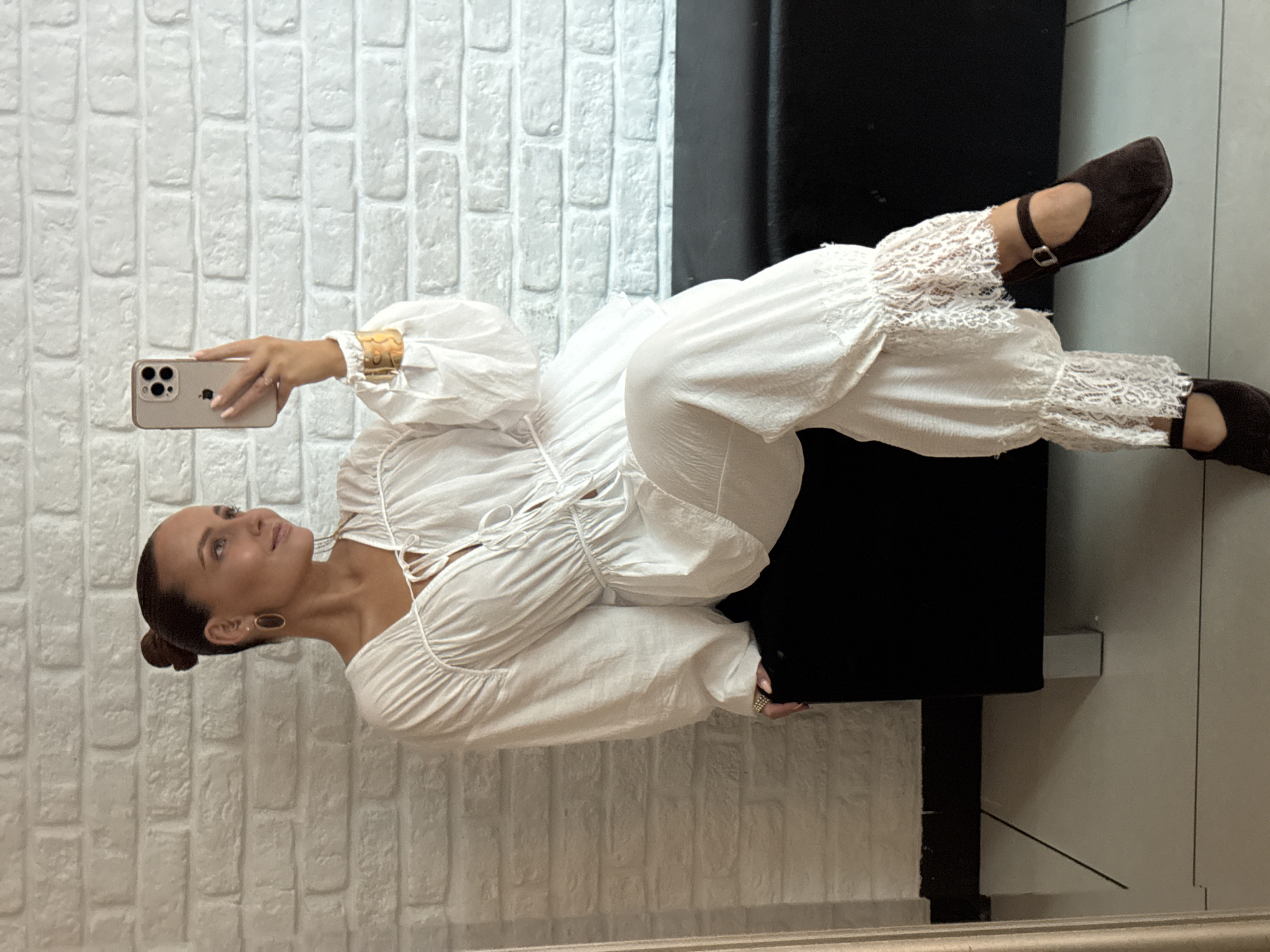 A woman is taking a selfie in a room with white brick walls and a black bench. She is wearing a white ruffled top and white pants with lace trim. She has her hair in a bun and is wearing gold earrings and a gold bracelet. Her phone is a gold iPhone. The room appears to be indoors and the lighting is natural. The woman is sitting on the black bench. The floor is tiled.