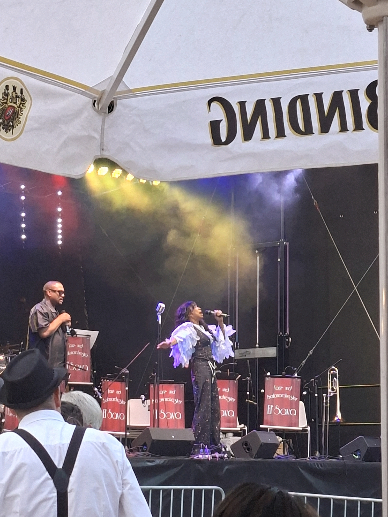 An outdoor stage event is depicted, likely in Frankfurt, Germany, given the presence of the BINDING logo prominently displayed on the tenting. The scene captures a musical performance with a female singer in the center, holding a microphone and wearing a sparkling dark outfit with ruffled sleeves. To her left stands a male performer, wearing sunglasses and a short-sleeved collared shirt, also holding a microphone. The stage is set up with various musical instruments, including drums, keyboards, and a trombone, some of which are decorated with red fabric bearing the "Loser und Balarechta El'Sava" text. Lighting from above casts colored glows and creates smoke effects on stage. In the foreground, the back of a man wearing a white shirt and suspenders, and a black hat is visible, suggesting an audience member. The overall ambiance suggests a lively concert or festival event.