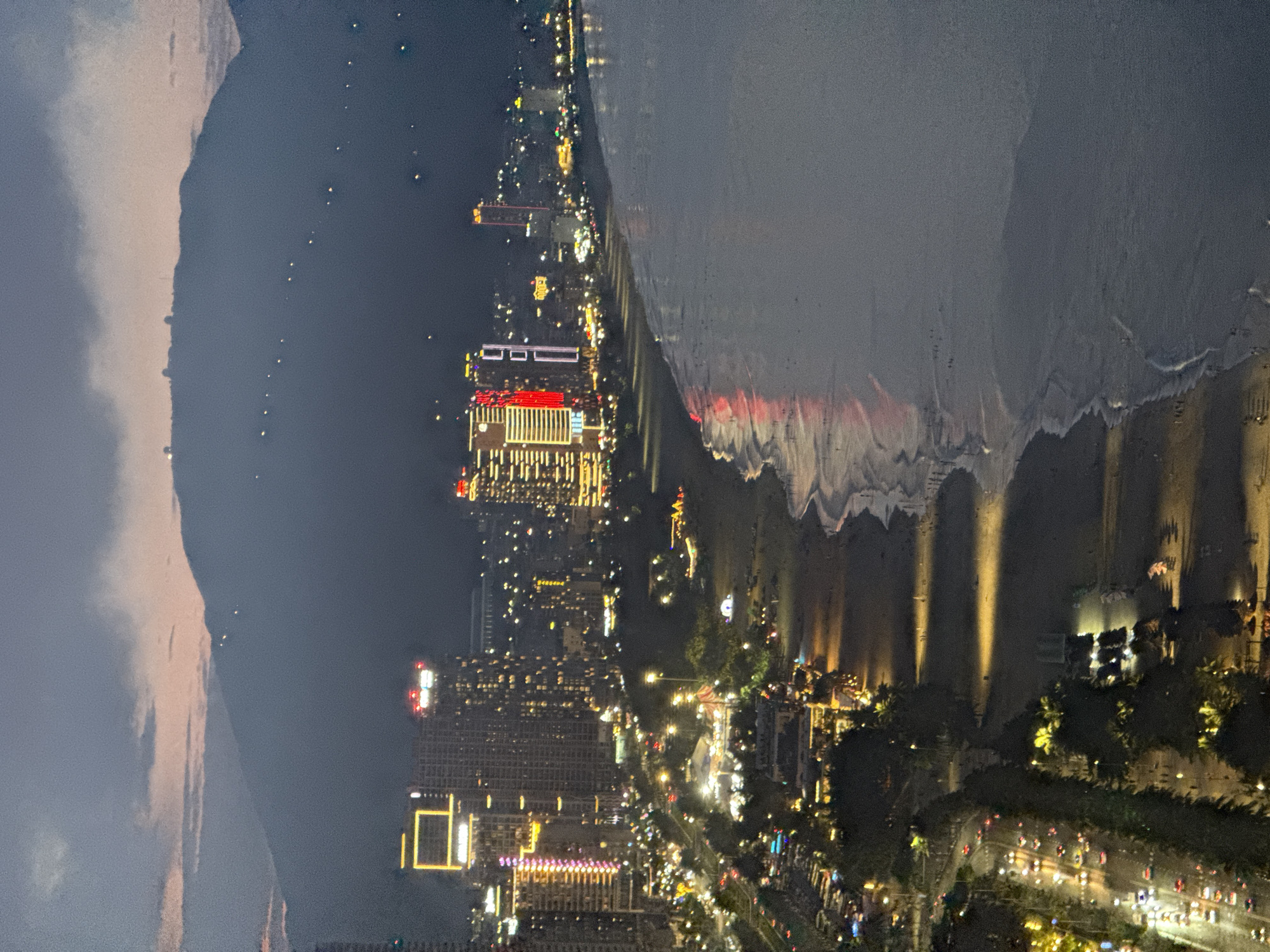The image depicts a night view of a coastal city, likely Đà Nẵng, Vietnam, given the context. The scene is dominated by a city skyline illuminated by numerous lights, stretching along a coastline. Tall buildings with brightly lit windows and facades are prominent. Some buildings feature striking red and yellow lights, suggesting hotels, commercial establishments, or entertainment venues.
Adjacent to the city is a body of water, possibly the ocean, with gentle waves lapping at the shore. The water reflects the city lights, creating a shimmering effect on the wet sand. The beach area appears to be lined with trees, some of which are also illuminated by streetlights or decorative lights, creating a warm, inviting atmosphere. A road or promenade runs along the coast, with traffic lights and car headlights visible, indicating a busy urban environment even at night.
The sky is dark, suggesting it is late evening or night. There are some clouds visible on the left side of the image, tinged with pink and purple hues, possibly from the last light of sunset or artificial lighting. The overall mood is one of vibrant urban nightlife and coastal beauty. No visible text is present in the image. The perspective is from a high vantage point, looking down on the city and coastline.