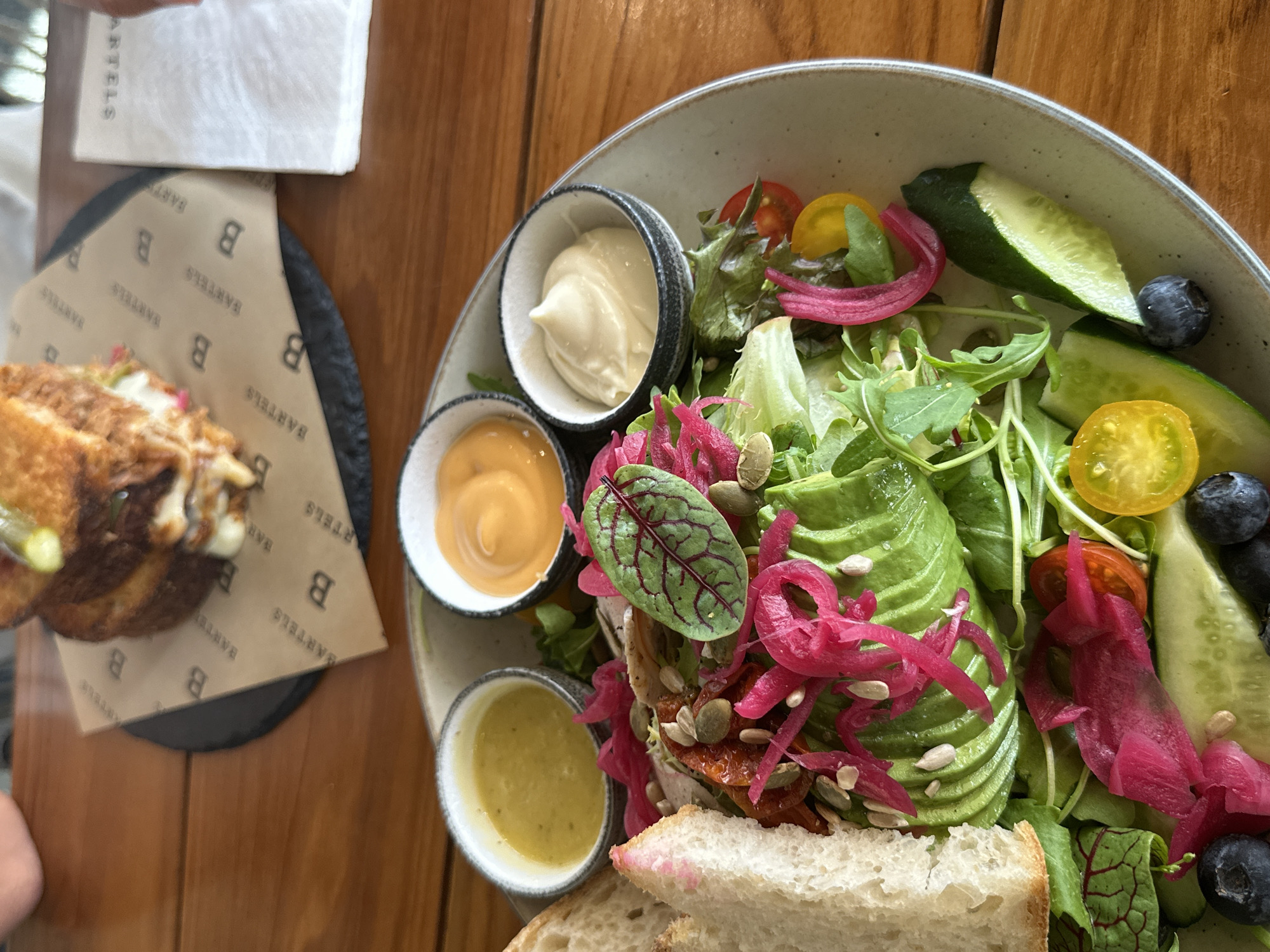 The image shows a plate of food, likely taken at a restaurant in Choeng Thale, Thailand, given the context. The food consists of a salad with avocado, pickled red onions, lettuce, tomatoes, cucumber, and blueberries. There are three small bowls containing sauces or dressings arranged alongside the salad. In the upper left corner of the image, there is a sandwich on a branded paper mat, along with a folded napkin. The food is sitting on a wooden table.