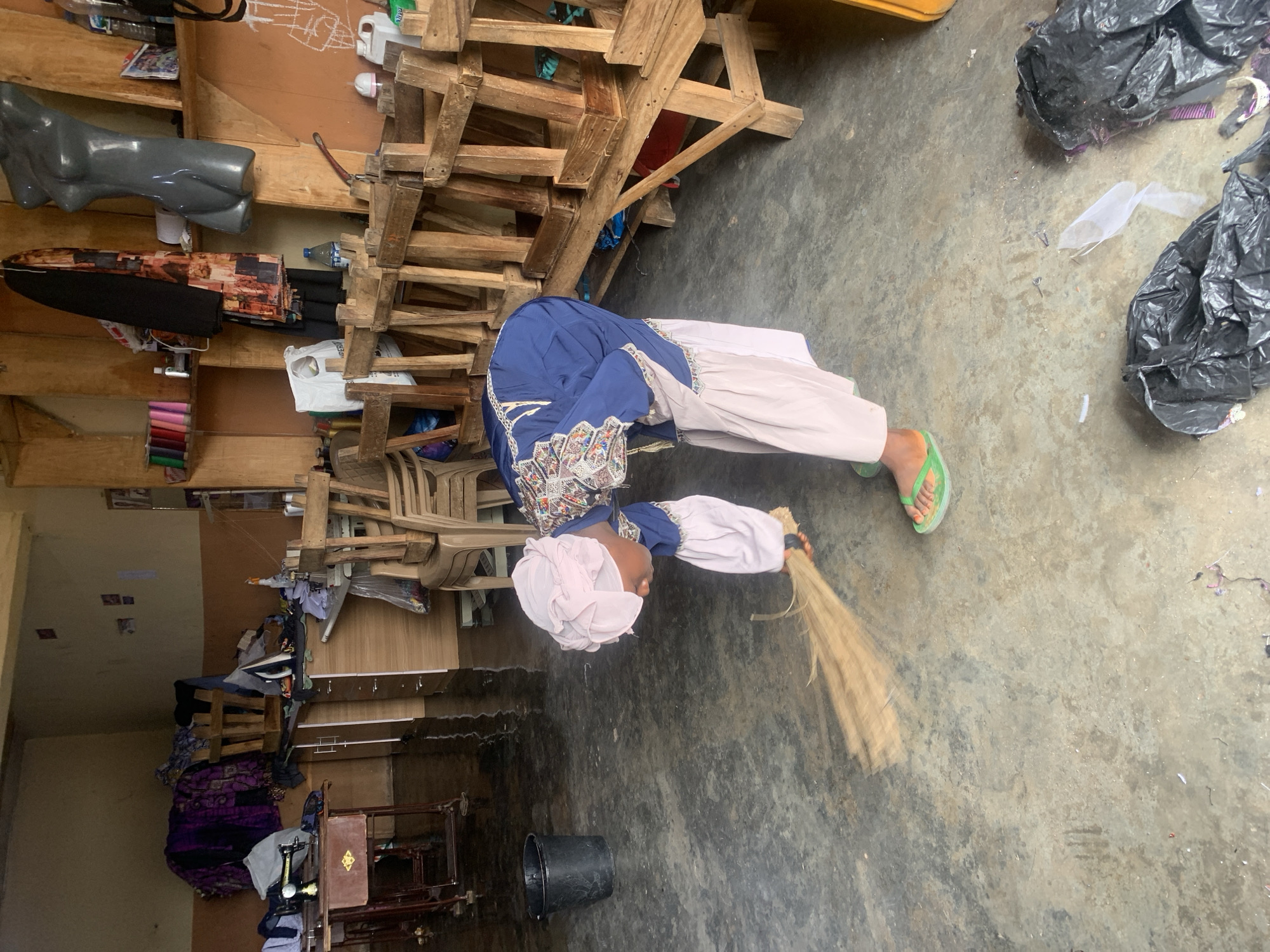 The image shows a person, likely a child, in a workshop or storage area. The person is wearing a blue and white traditional outfit with a pink headscarf. They are bent over, holding a broom and appear to be sweeping the concrete floor. A black bucket is visible on the floor near them. The setting is cluttered with various items, including wooden shelves stacked with fabrics, mannequins, and discarded black plastic bags. In the background, there are more shelves filled with what appears to be chairs and other materials. The lighting suggests it could be daytime. The overall impression is one of a functional, perhaps slightly disorganized, workspace.