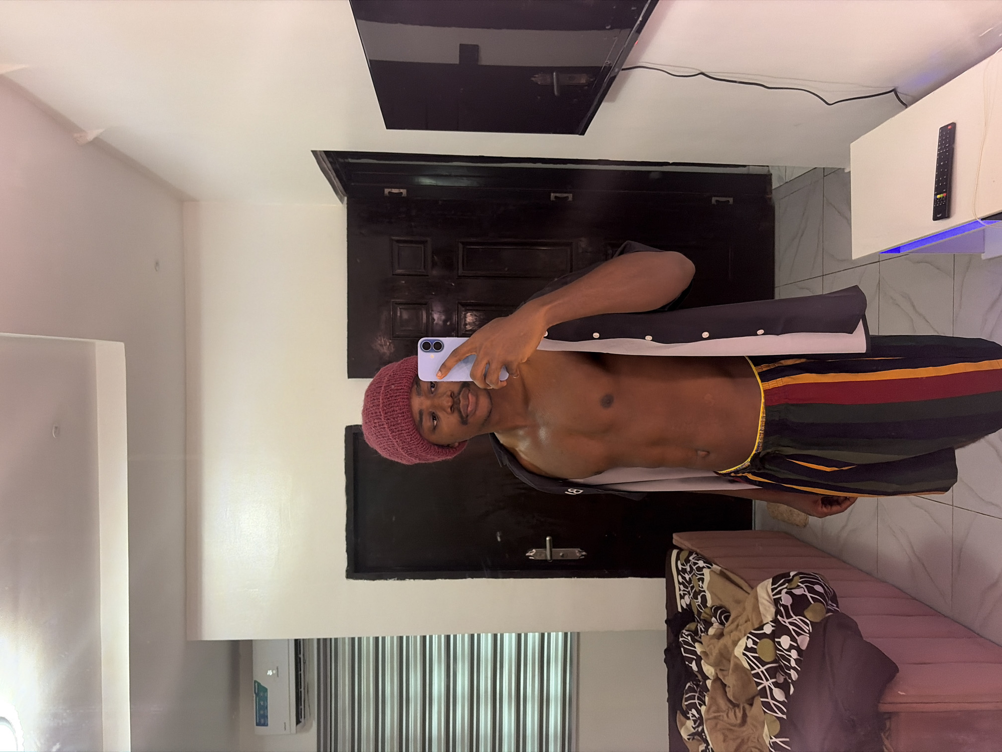 A male with brown skin, a maroon beanie, bare chest, and striped shorts is taking a selfie with a light purple smartphone in what appears to be a residential interior in Igboefon, Nigeria. He is standing in front of a dark brown door, likely a bedroom door. A flat-screen television is mounted on the wall above the door. A white cabinet with a remote control on top is visible to the upper right. A narrow bench with folded clothing is present in the lower right corner. White Venetian blinds cover a window at the bottom of the frame. There is an air conditioner visible on the wall to the left. The walls are painted white, and the floor appears to be tiled.