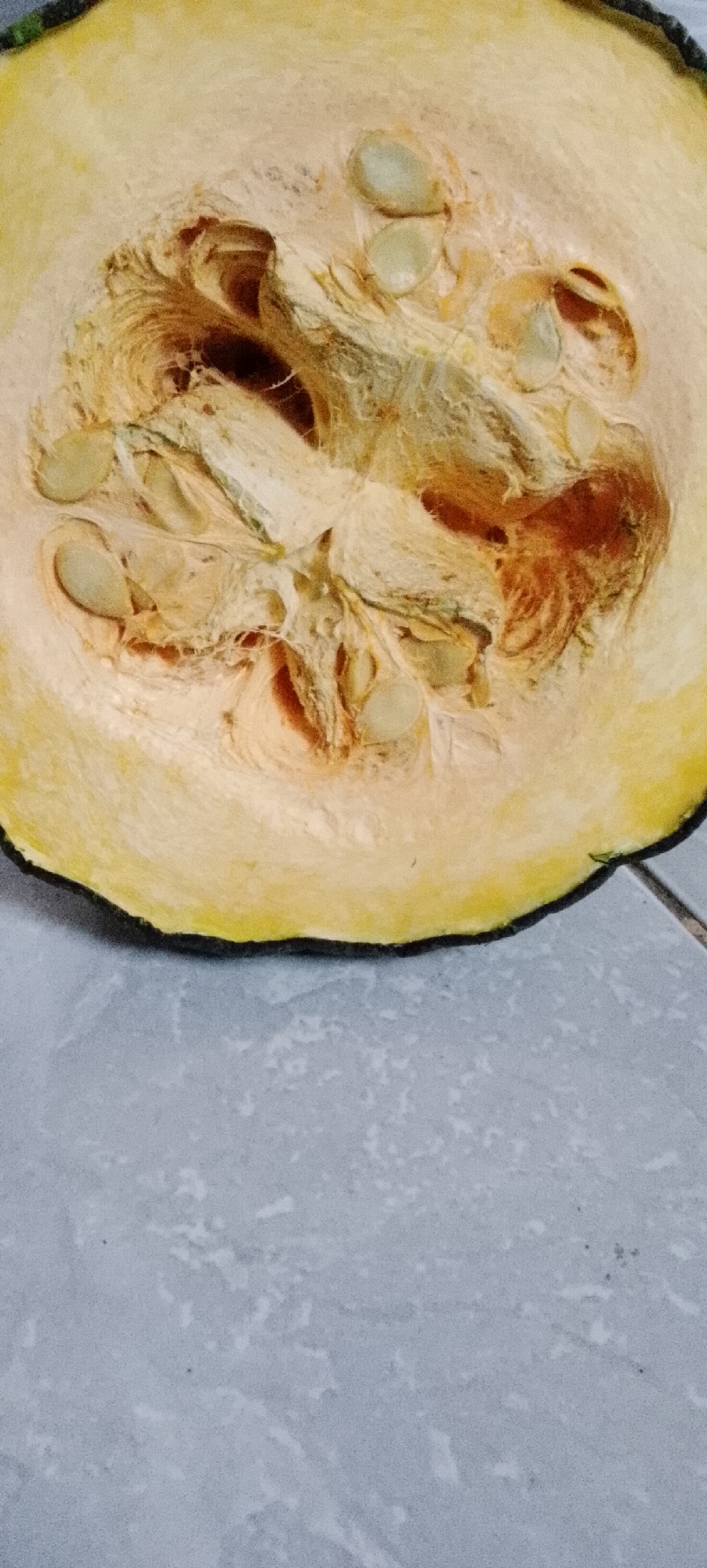 The image shows a close-up, overhead view of a cut pumpkin. The pumpkin is a pale yellow on the inside with visible seeds and stringy pulp. The outer rind is a dark green. The pumpkin is resting on a grey tiled surface. The image appears to have been taken indoors. There is no information in the image to suggest a location in Padang Dalom, Indonesia.