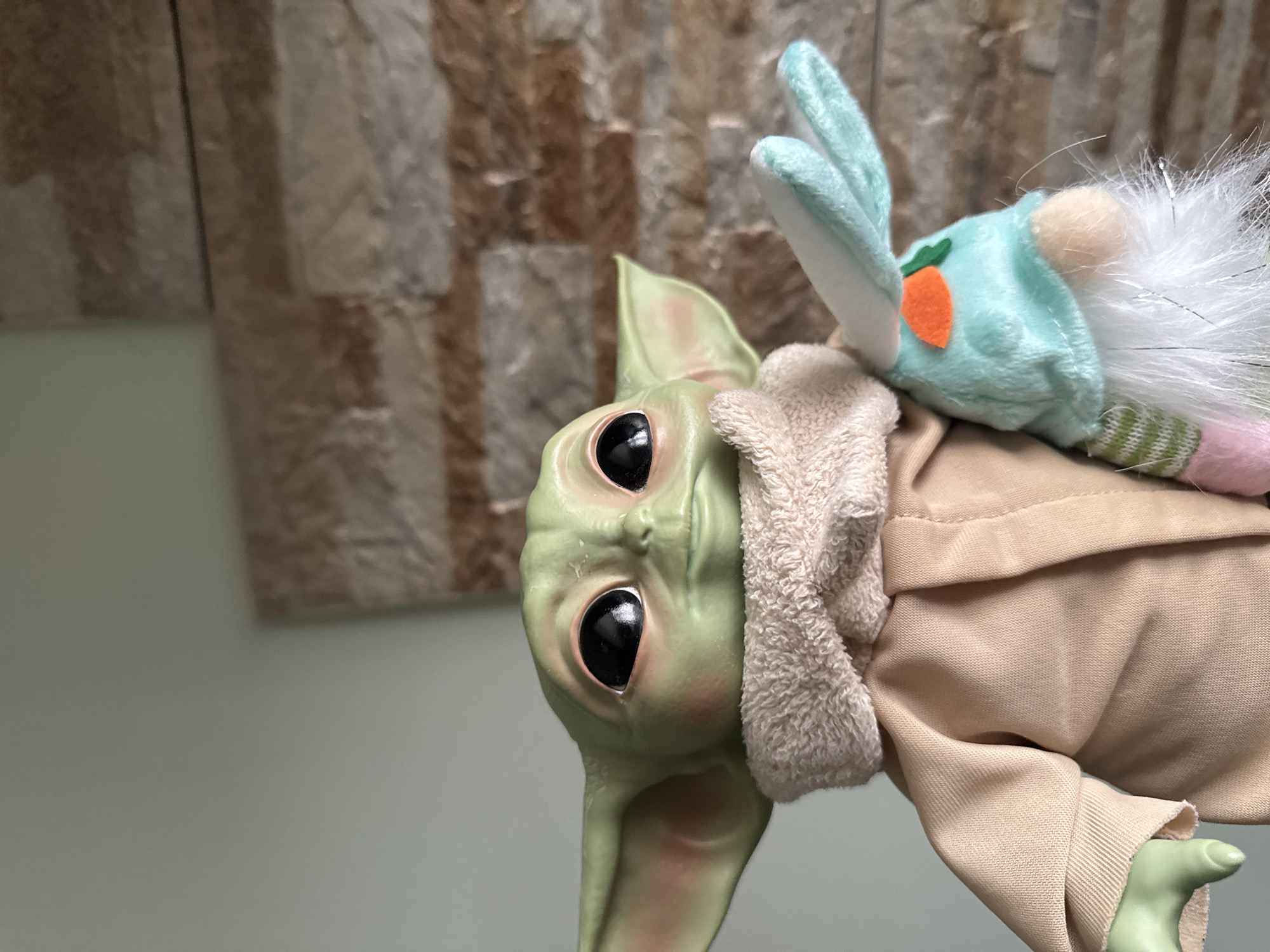 A close-up shot shows a Grogu doll wearing a brown robe with a light tan collar. A stuffed rabbit toy is placed on top of the doll's head. The background consists of a vertically-aligned textured wall on the right, and a light grey wall to the left.