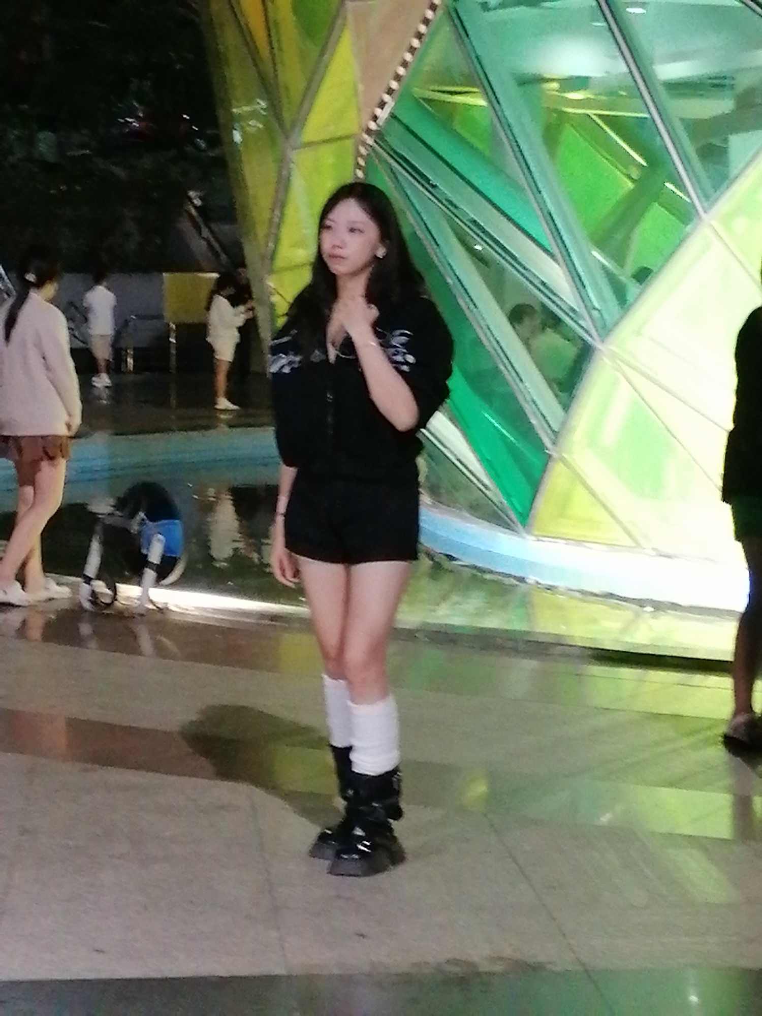 The image shows a young woman standing in front of a large, geometric structure with green and yellow faceted glass panels. She is wearing a black zip-up jacket, black shorts, white knee-high socks, and chunky black platform boots. Her hair is long and dark, and she is looking slightly off to the side with her hand near her chin. Several other people are visible in the background, some appearing to be walking or standing. To the left, a person in a light-colored jacket and a brown skirt is walking away from the camera. Further in the background, another person in a white shirt and shorts is visible. The ground appears to be wet, reflecting the light from the structure and the surroundings. A partially visible blue object, possibly part of a water feature or a piece of equipment, is on the wet ground to the left. The lighting suggests it might be evening or nighttime, with artificial lights illuminating the scene. The overall setting appears to be a public space, possibly a park, plaza, or an area with an architectural installation. The geometric structure with its vibrant colors is a prominent feature of the scene.