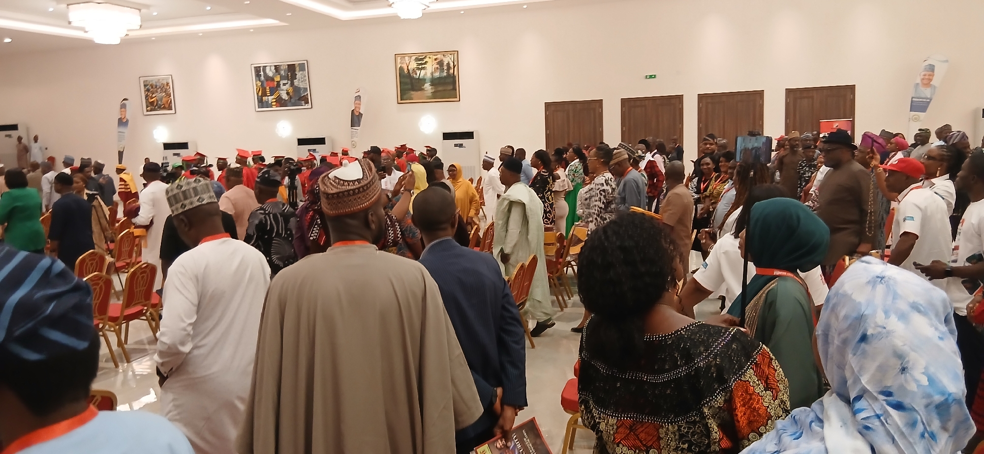 The image depicts a large indoor gathering in a well-lit room, likely a ballroom or convention hall in Unknown, Unknown. A crowd of people, mostly dressed in formal or semi-formal attire, fills the space. Many individuals are wearing traditional African clothing, including robes, headwraps, and caps. There is a mix of ages and genders present.
Several people are moving about, suggesting a networking event or pre-function mingling. A few are holding phones, possibly taking pictures or videos. Rows of empty red chairs line one side of the room.
The room's interior features white walls decorated with framed artwork and tall, dark brown doors. A large chandelier hangs from the ceiling. There are air conditioning units on the wall. There is a promotional banner in the corner.