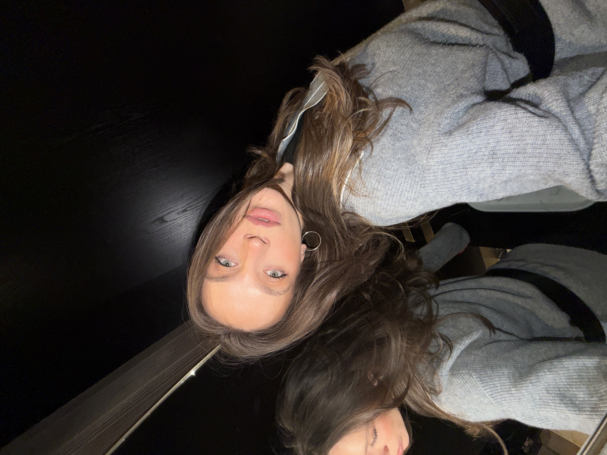 The image is an upside-down selfie taken of a woman with light eyes and brown hair, wearing a gray sweater. The background is a dark, glossy surface, likely a table, which creates a partial reflection of the woman. A portion of what may be a white collared shirt is visible beneath her sweater. The location in the city of Budva, Montenegro, cannot be determined from the image.