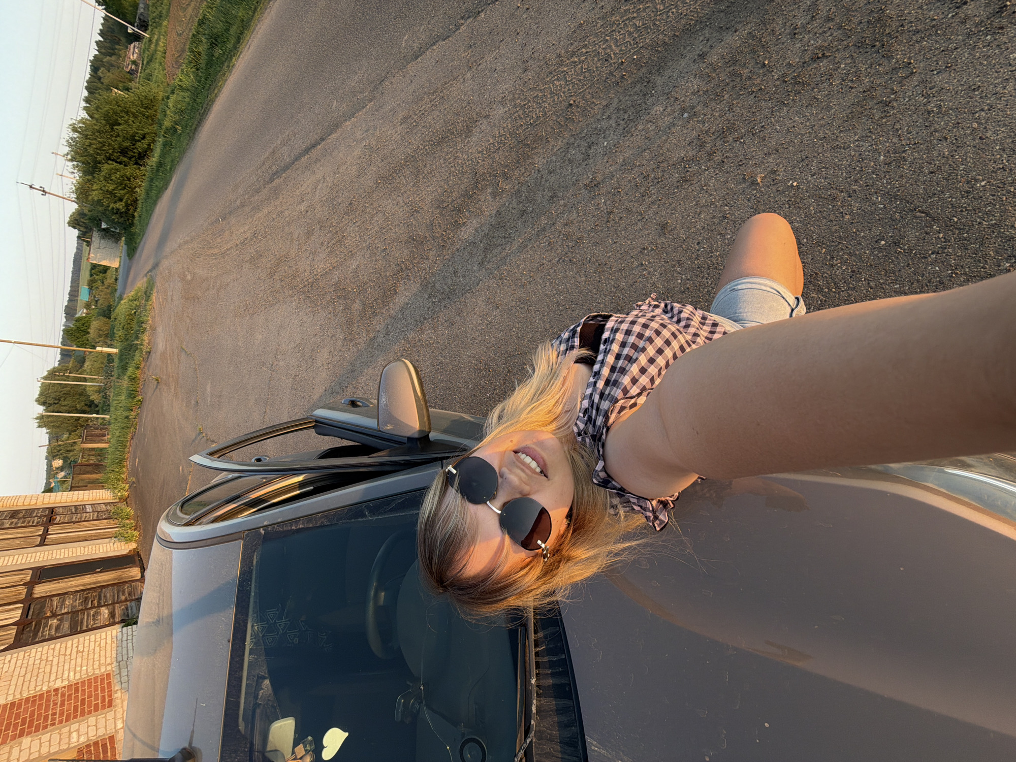 A young woman is posing for a selfie while lying on the hood of a gray car. She is wearing sunglasses and a checkered shirt, with light-colored hair. The car door is open. The scene appears to be on a road in the outskirts of a settlement. Based on the information provided, the location is Чумна, Russia. Adjacent to the road are grassy areas and buildings. The photo is taken at ground level.