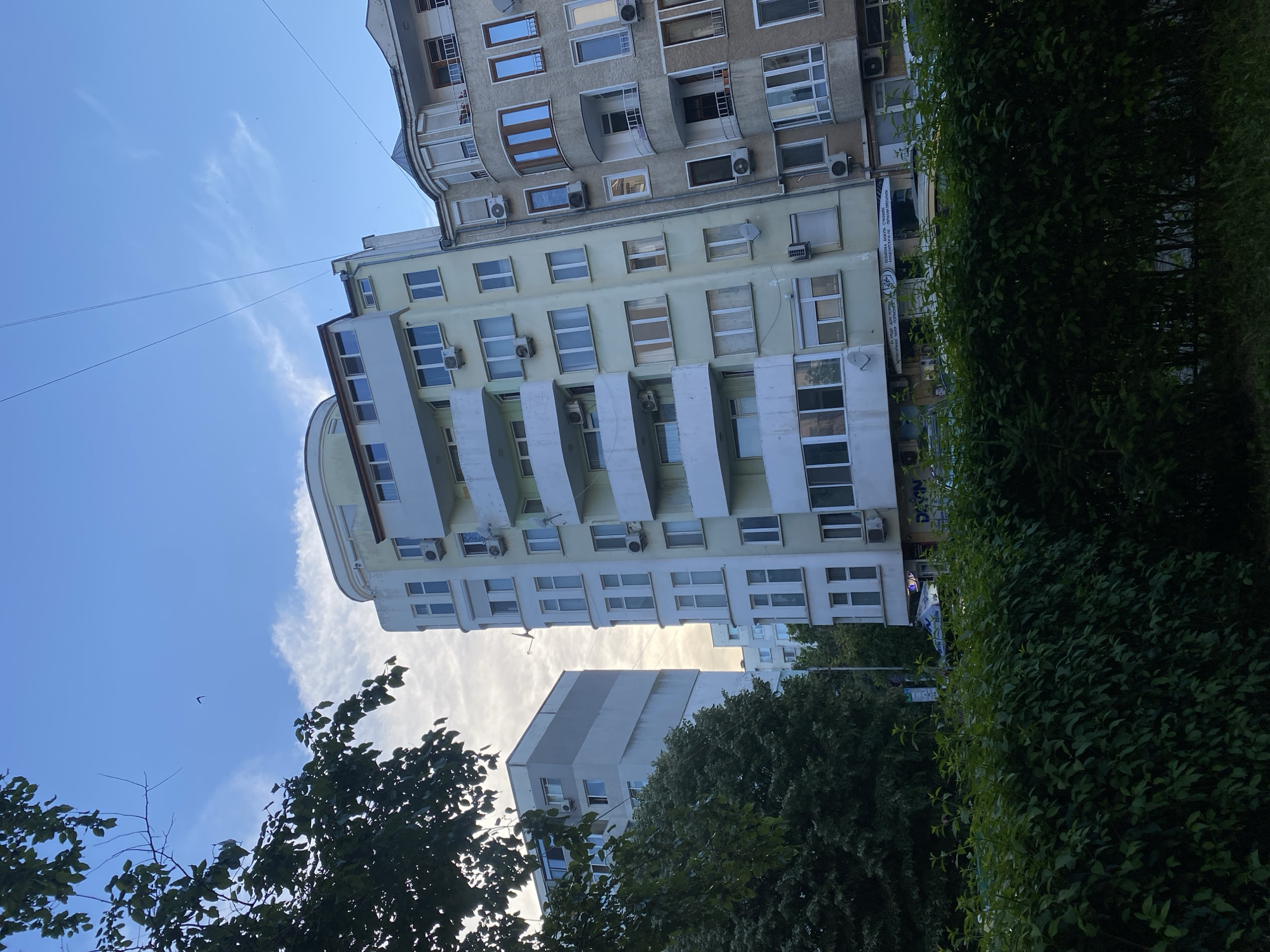 The image shows a street scene in Varna, Bulgaria. The foreground is dominated by a dense, well-maintained hedge. Behind the hedge, several multi-story apartment buildings are visible. The buildings exhibit a mix of architectural styles and colors, with some featuring light yellow facades and others a more muted grey. Balconies with white railings are prominent on some structures. Several windows, some with closed blinds, are scattered across the building facades, along with air conditioning units mounted on the exteriors. A few trees with dark green foliage are interspersed between the buildings and the hedge. The sky above is a light blue, with scattered white clouds and what appear to be power lines stretching across the frame. A small bird is visible in flight against the sky. The overall impression is of a residential area in a city.