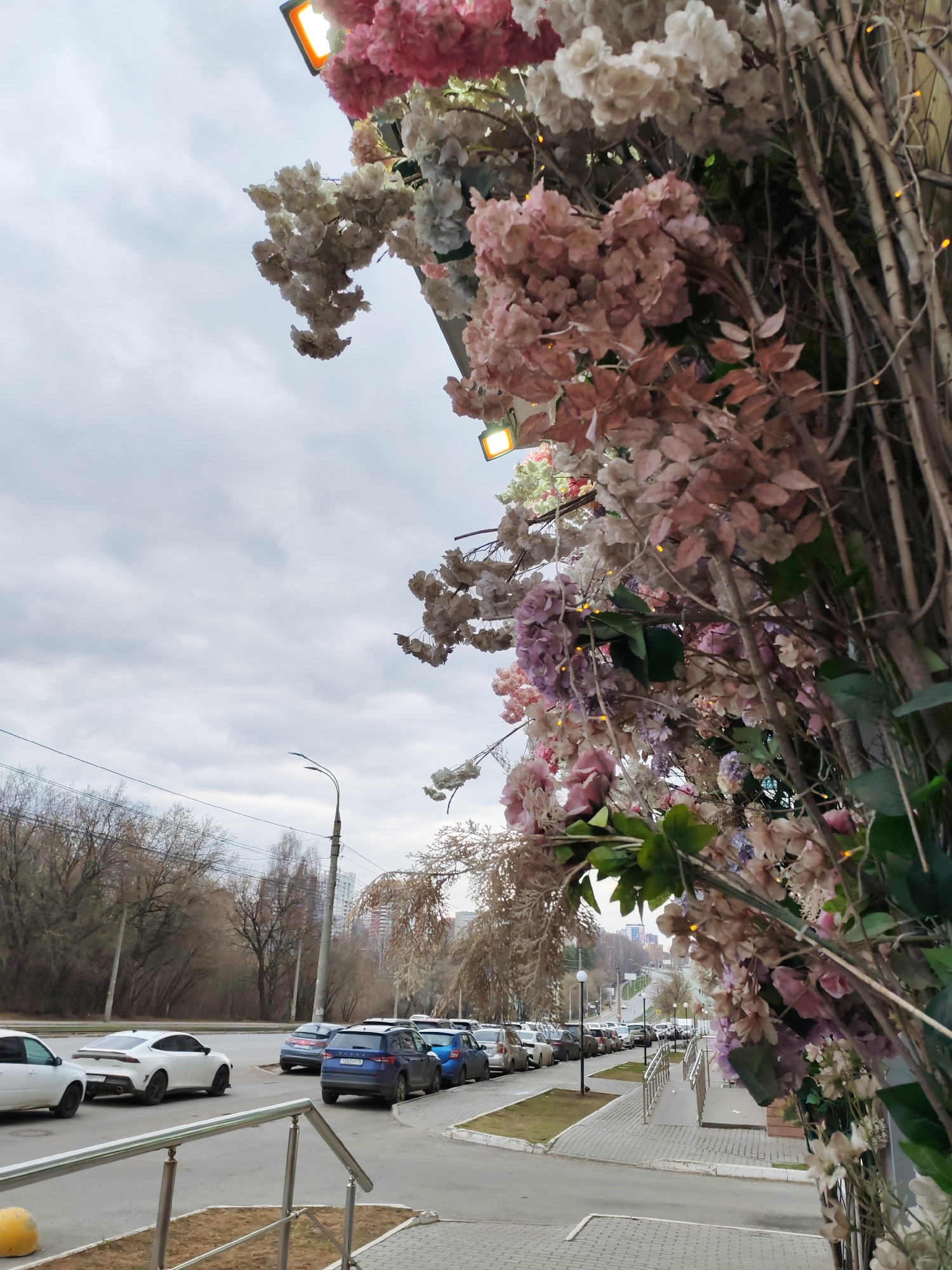 The image shows an outdoor scene in Izhevsk, Russia, under an overcast sky. The foreground is decorated with an archway of artificial flowers in shades of pink, white, and purple, intertwined with bare branches and subtle string lights. Adjacent to the archway is a pedestrian walkway with a metal railing, leading to a street lined with parked cars. On the left side of the street, bare trees are visible, suggesting it might be early spring or late autumn. In the background, there are buildings of varying heights, indicating an urban setting. Streetlights are also visible along the street. The overall impression is of a decorated outdoor space next to a typical city street in Izhevsk.