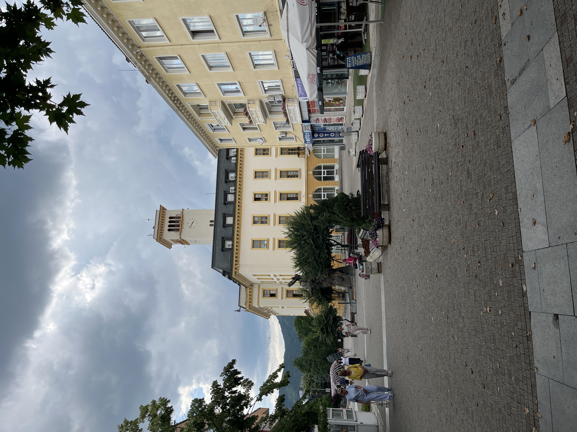 The image depicts a street scene in Sliven, Bulgaria, likely in the central pedestrian zone near the Clock Tower. In the foreground, a paved pedestrian walkway is visible, with scattered fallen leaves suggesting an autumn season. Several people are walking on the walkway. To the right, there is an outdoor seating area under a large umbrella with tables and chairs, and a kiosk selling beverages, with visible branding for Coca-Cola and Pepsi. Adjacent to this are storefronts with awnings. Dominating the background is a large, pale yellow building with a prominent clock tower. This building has multiple windows and architectural details. To the right of this building is another, taller pale yellow building with balconies and numerous windows. Green trees and foliage are present throughout the scene, and a range of hills can be seen in the distance under a cloudy sky.