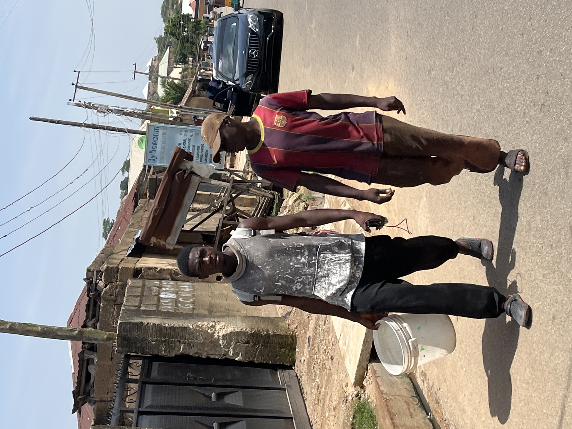 Two young Black males are walking on a paved road in Jos, Nigeria. The male on the left is carrying a white bucket in his right hand and a phone in his left. He is wearing a gray sleeveless shirt with white splatters, black pants, and gray sneakers. He has a backpack on his shoulders. The male on the right is wearing a maroon and purple striped t-shirt, brown pants, and dark sandals. He is wearing a brown baseball cap. Behind them, there is a black Mercedes-Benz car and buildings in the background. The buildings appear to be made of brick and corrugated metal. The sky is blue with some clouds. The scene depicts a street in a residential or commercial area of Jos.