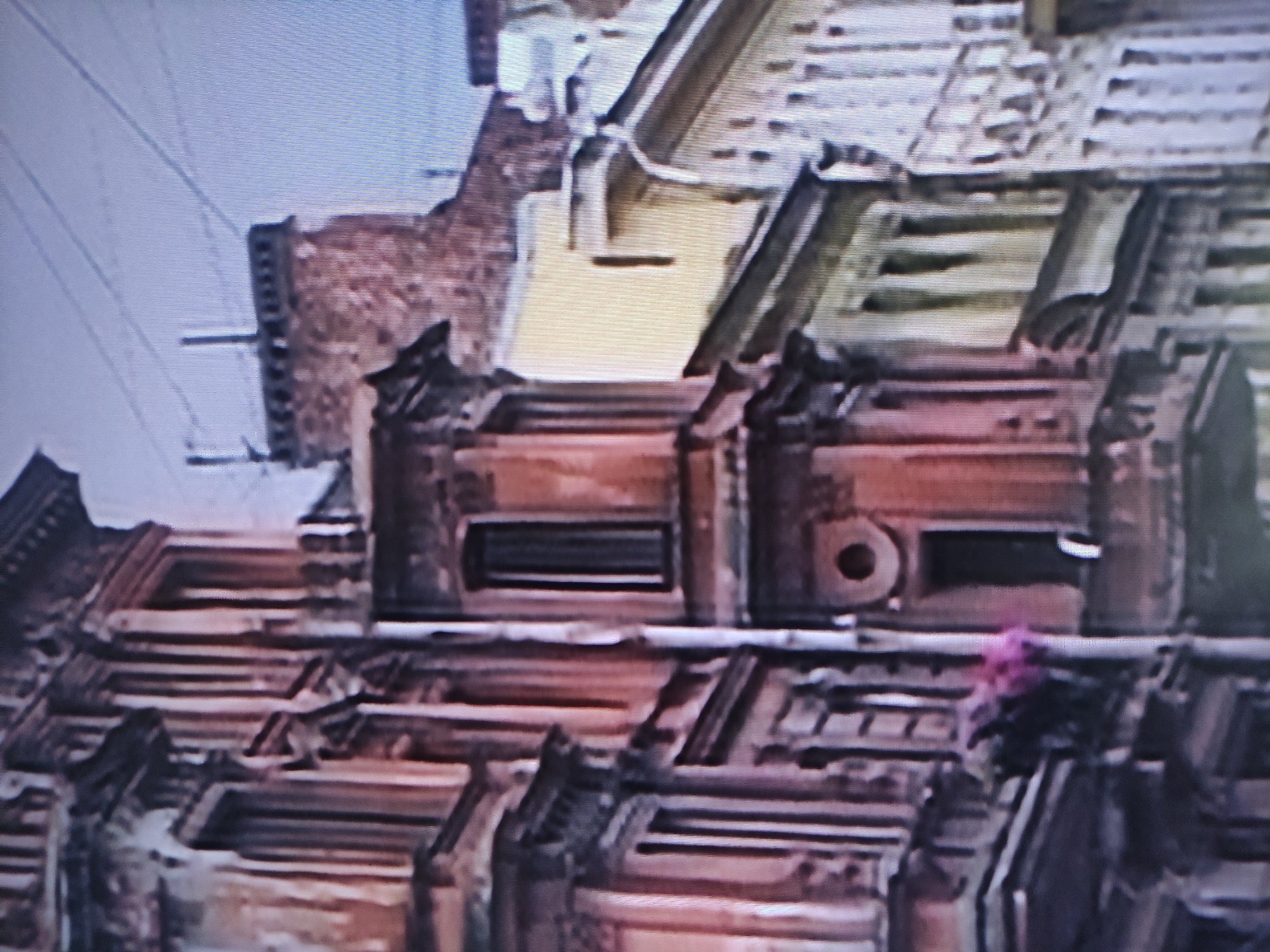 This low-resolution, blurry image, likely a still from an older video, presents an elevated view of an industrial or urban landscape during daytime, rotated clockwise. The scene is dominated by complex building structures, primarily rooftops and upper facades, under a clear, light blue sky visible in the upper-left quadrant.
The main subjects are reddish-brown brick or concrete buildings with intricate architectural details. In the midground, a distinct structure features a flat, yellowish-beige roof section with a dark, rectangular opening or vent. Adjacent to it on the right, another similar structure shows a circular opening and a rectangular slot, suggesting industrial machinery or large ventilation components.
Further down, the structures exhibit layered and stepped rooflines with various geometric shapes, possibly indicating multiple levels, skylights, or specialized industrial construction. Some roof sections appear corrugated or segmented.
The pale blue sky in the upper left is traversed by several thin, dark lines, which are likely power lines or cables. There are no people visible, and no discernible activity is taking place; the scene appears static. A small, blurry, bright pink or purple object, possibly a piece of debris or fabric, is visible on one of the structures in the lower-right portion of the image.
The image quality is characterized by visible horizontal scan lines or interlacing artifacts. No readable text is present in the image. The industrial architecture is consistent with an urban area such as Pavlodar, Kazakhstan.