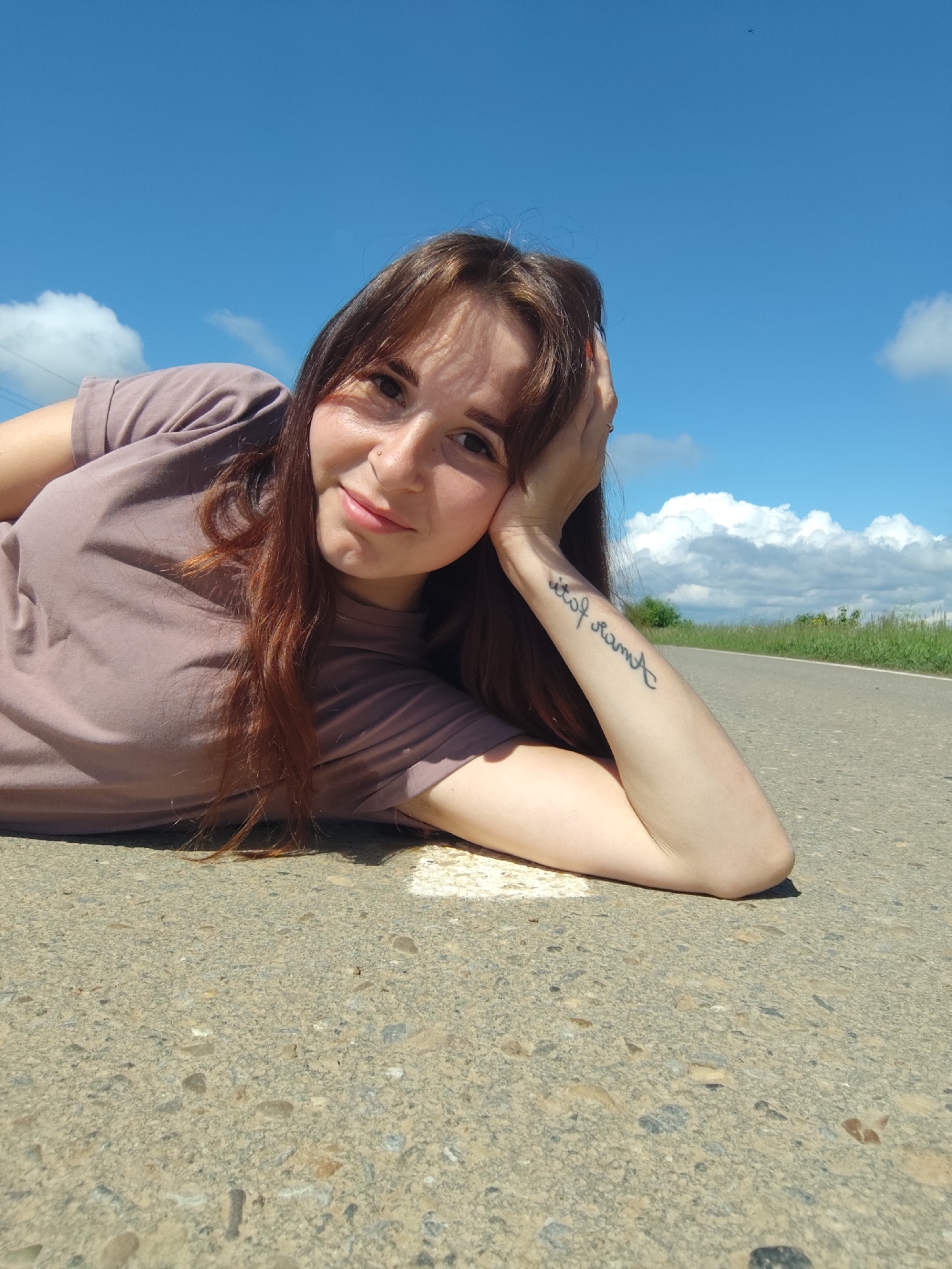 A young woman is lying on her side on an asphalt road in Фоки, Russia, posing for a photograph. She has long, reddish-brown hair and is wearing a light purple short-sleeved t-shirt. Her head is resting on her left hand, with her left elbow propping her up. She is looking directly at the camera with a calm, pleasant expression and a slight smile. A small piercing is visible on her nose.
The scene is outdoors, under a clear, bright blue sky with scattered white, fluffy clouds, indicating a sunny daytime. The camera is positioned low, capturing a wide expanse of the sky above her. The road surface beneath her is asphalt with visible aggregate. To her right, the road stretches into the background, flanked by a grassy verge and distant green foliage and trees.
A notable detail is a black cursive tattoo on her left forearm, which appears to read "out of reach" when viewed from her perspective. The overall impression is one of leisure and serenity in a rural or semi-rural setting.