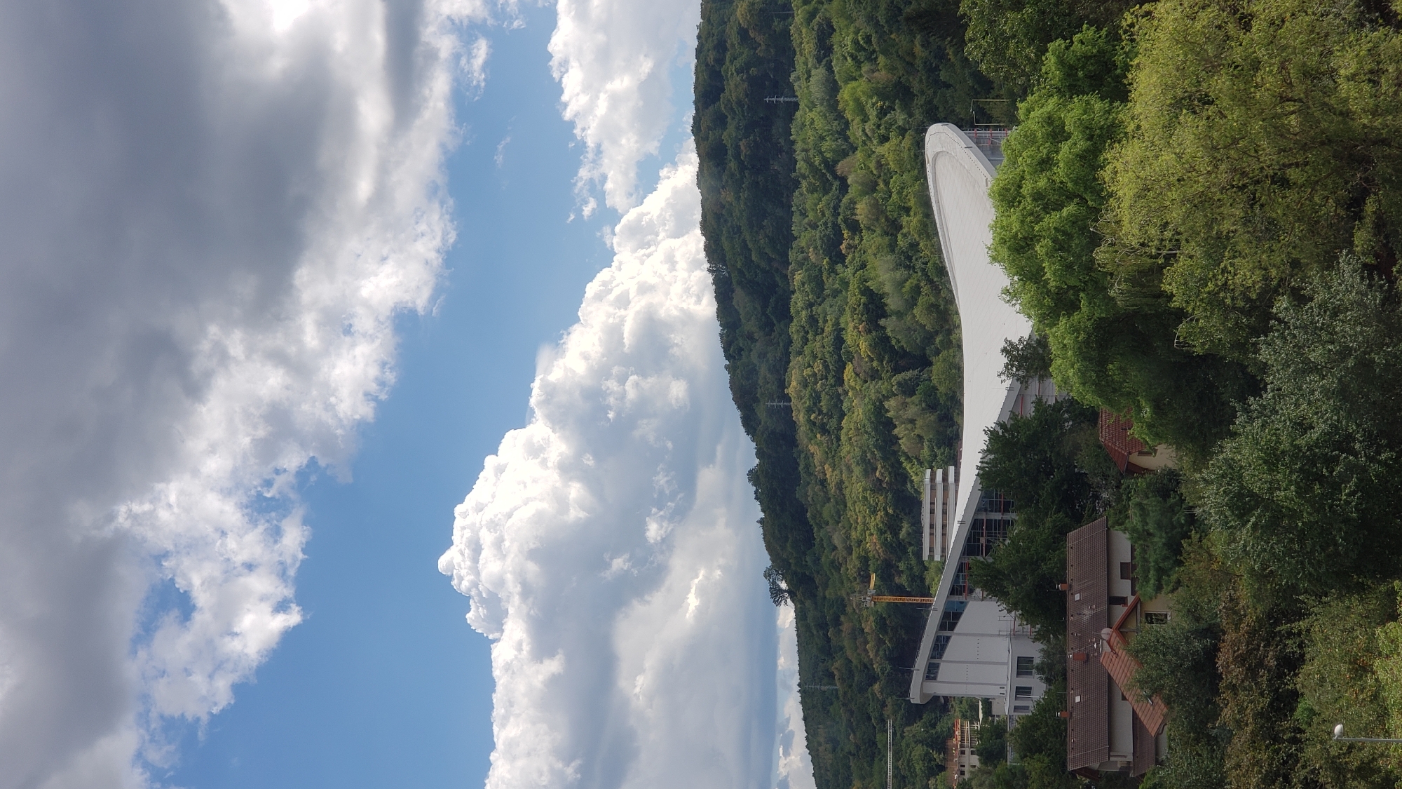 The image captures a view of the city of Prešov, Slovakia, dominated by a lush, green hillside covered in dense forest. In the foreground, several residential buildings with red tile roofs are nestled amongst the trees. A large, modern building with a distinctive curved white roof and glass facade is situated behind the houses, with scaffolding visible on one side, suggesting ongoing construction or renovation. A yellow crane is also present near this structure. The sky above is a dramatic mix of bright blue and heavy, dark gray clouds, with fluffy white clouds scattered throughout. The overall impression is a suburban or semi-rural landscape within the city.