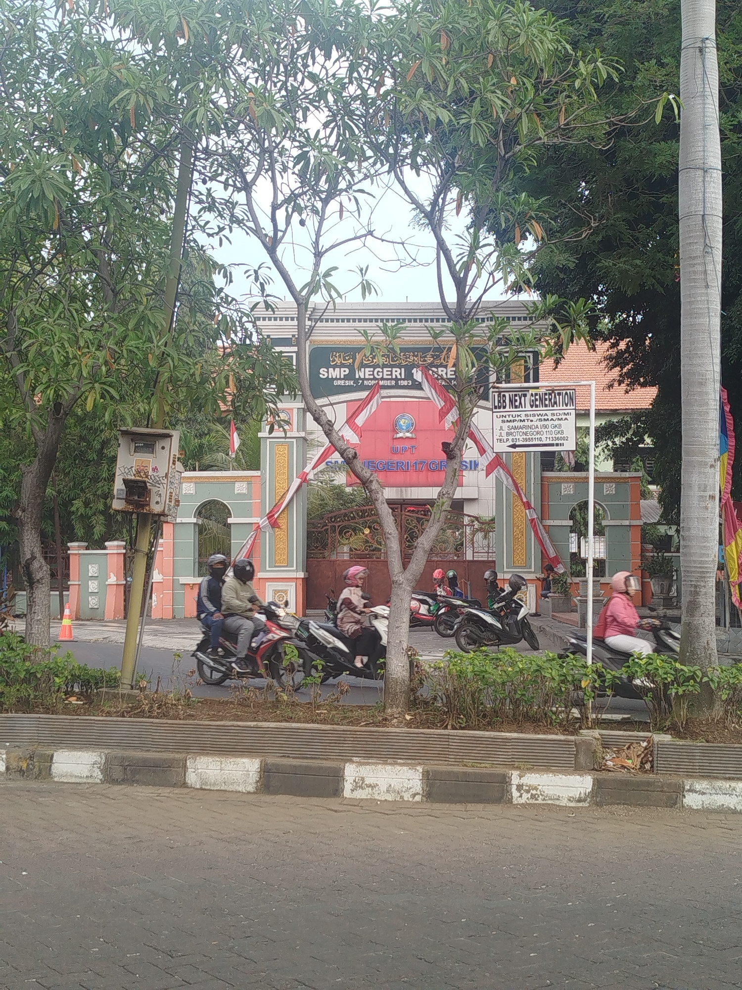 The image depicts the entrance to SMP Negeri 1 Gresik, a public junior high school in Gresik, East Java, Indonesia. The school gate is adorned with red and white Indonesian flags. Several students on motorcycles are seen in the foreground, suggesting a typical school day. The environment is urban, with a paved road and sidewalk. A sign for "LBB NEXT GENERATION" is visible, indicating a tutoring or educational service nearby. Trees and vegetation surround the school, contributing to a leafy, green atmosphere. The overall scene conveys a sense of everyday life and education within an Indonesian city.