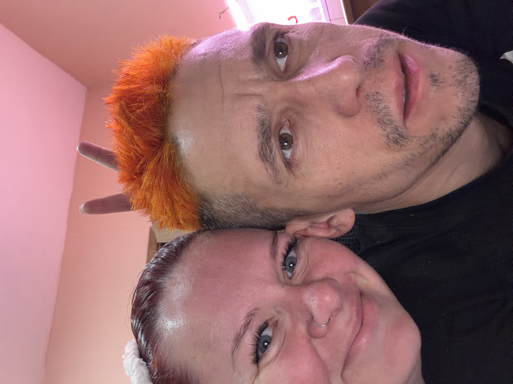 Two individuals are posed closely for a selfie. The person on top has short, spiky orange hair and a light complexion with stubble. The person underneath has reddish-brown hair and a fair complexion, with a nose ring visible. The background is a pale pink color. A pair of ears are sticking out the top of the man’s head. The image lacks any explicit references to Chaikovsky, Russia, or any indications of a specific location.
