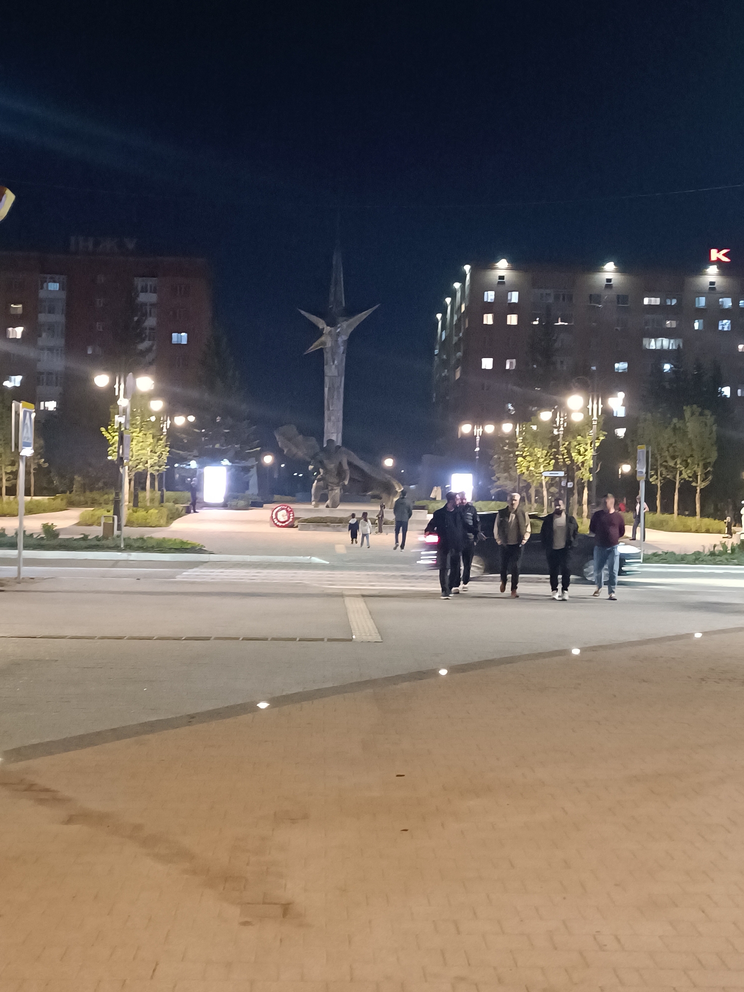 The image depicts a night scene in Kokshetau, Kazakhstan, featuring a street intersection with pedestrians crossing.
Environment: It is nighttime with visible artificial lighting from street lamps and buildings. The ground is paved. Buildings are visible in the background, including one with the Cyrillic letters "IHKV" on top and another with a red Cyrillic letter "K".
Objects: A tall monument with a star-like structure on top dominates the center background. There are traffic signs and what appears to be a memorial wreath. A dark-colored vehicle is parked near the pedestrian crossing.
People: Several individuals are walking across the pedestrian crossing. A group of five men are notably visible, walking in the foreground. Other people are seen in the background, including children.
Actions/Interactions: People are walking across the street. There are no visible interactions or activities other than walking.