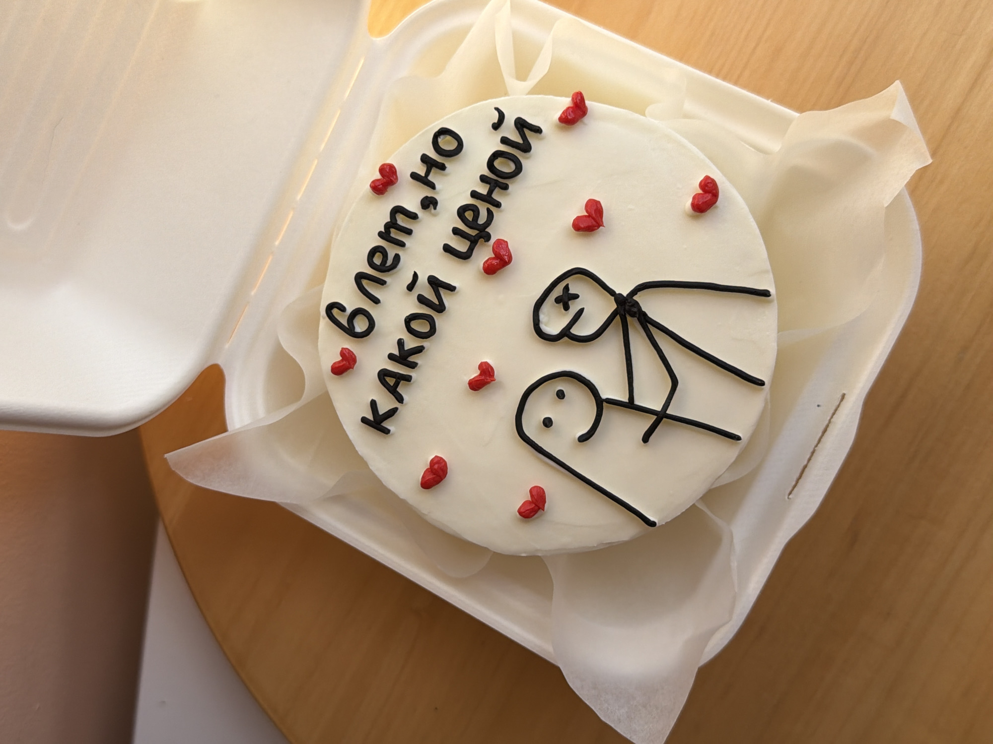 A round, white-frosted cake is centrally positioned within an open, white, disposable clamshell container, lined with translucent white paper. The cake features black icing lettering in Cyrillic, spelling "6 лет, но КАКОЙ ЦЕНОЙ" (translating to "6 years, but AT WHAT COST"), and two minimalist stick figures. The left figure depicts a smiling individual with dot eyes, while the right figure has X-shaped eyes, suggesting distress, and is being held by the left figure. Approximately a dozen small, red heart-shaped adornments are scattered across the cake's surface. The container rests on a polished light brown wooden surface, possibly a table or countertop, with a blurred light-colored background. The scene is depicted in Tel-Aviv, Israel.