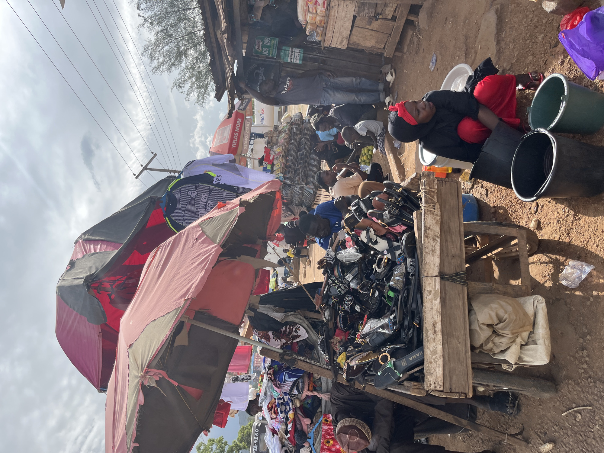 The image depicts a bustling outdoor market scene in Rafin Par, Nigeria. The foreground is dominated by a wooden cart overflowing with various footwear, primarily sandals and flip-flops, of different styles and colors. To the left, a man in a dark brown hat and dark clothing is partially visible, appearing to be tending to the merchandise. Behind the cart, several people are gathered, some sitting and others standing.
Above the cart, two large market umbrellas, one pink and one a combination of pink and black, provide shade. Hanging from a line between the umbrellas are several pieces of clothing, including what appear to be sports jerseys with "Real Madrid" branding.
In the mid-ground, a makeshift stall is visible with a wooden roof and shelving. Several loaves of bread are stacked on the shelves. A man in a grey t-shirt and blue jeans stands near this stall, holding his arm up. Other individuals are seen interacting in this area, some seated on the ground.
To the right of the scene, a woman wearing a black headscarf and red clothing is crouched down next to several buckets, one black and one green. She is looking towards the left. A white basin is beside her. Various plastic bags are scattered on the ground around her.
The ground of the market appears to be dirt. In the background, there are more stalls and structures, as well as trees and a cloudy sky with power lines crisscrossing it. A vehicle with a license plate reading "PTA 4160" is visible in the far background on the left. The overall impression is one of a lively and active marketplace.