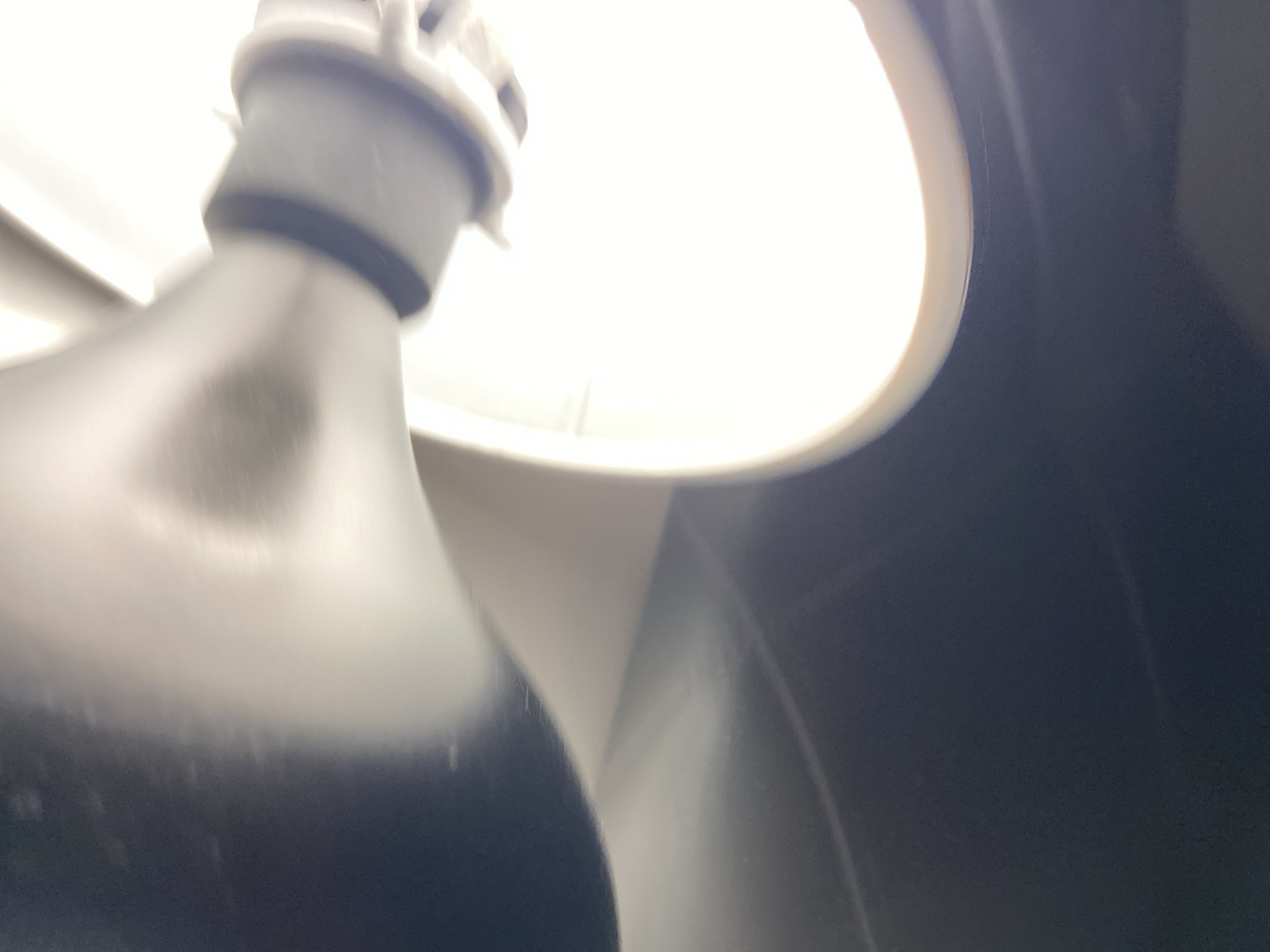 A close-up, upward-angled, and partially blurred image depicts an interior light fixture located in Budva, Montenegro. The primary subject is a bright, intensely luminous white light source, appearing as a semi-circular or oval shape in the upper right portion of the frame, emitting diffused light and exhibiting a thin, lighter-colored rim. Adjacent to this light, extending from the lower-left quadrant, is a dark, indistinct object. This object possesses an upper cylindrical section, light grey in color, detailed with a distinct black band and white protrusions at its uppermost part, consistent with the base of a bulb or fixture component. This cylindrical element transitions into a wider, darker, and somewhat bulbous body that fills the lower-left area. The background immediately surrounding the bright light is a clean, bright white surface, likely a ceiling, while the remaining background area, particularly to the right and lower-right, is dark and undifferentiated. No individuals, specific actions, or additional objects are discernible within the frame. The scene represents an illuminated interior environment.