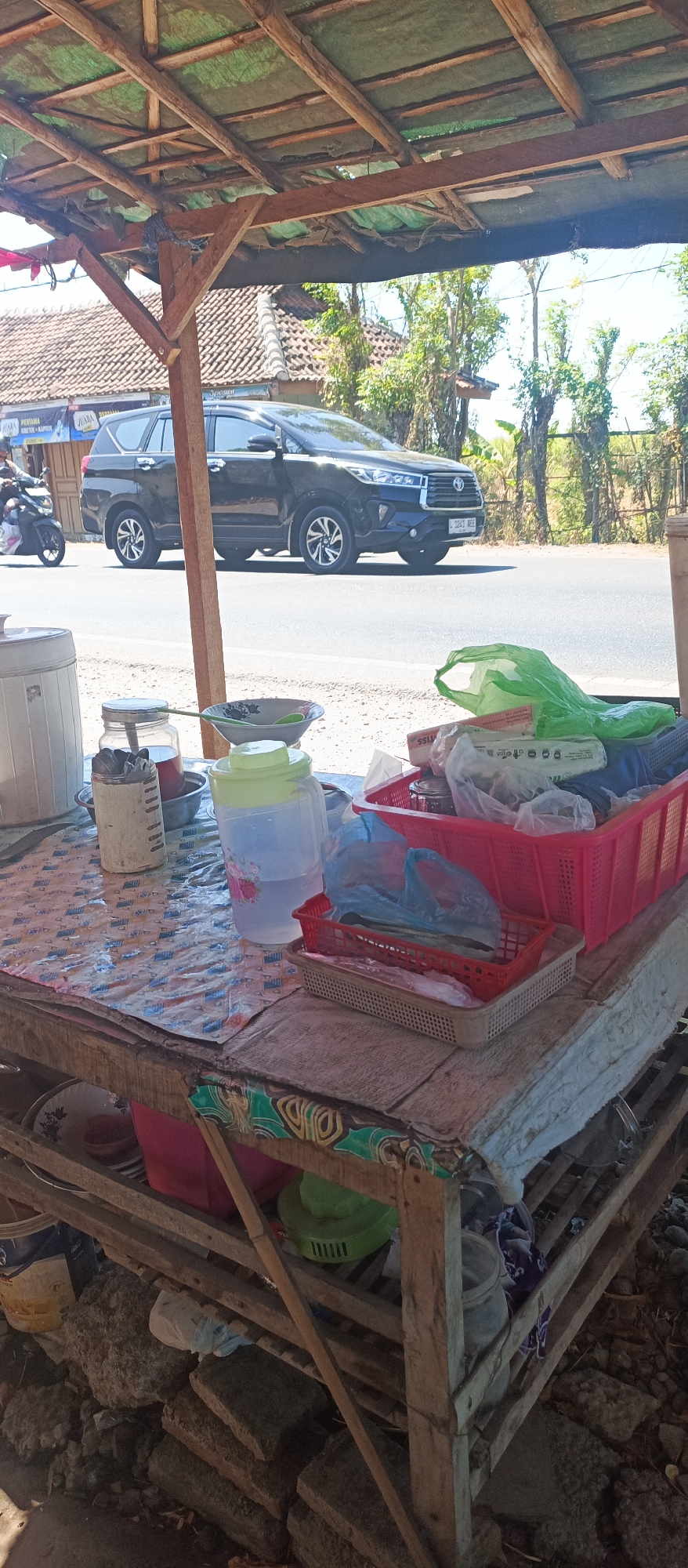 An outdoor food stall is situated beside a paved road in Jatisari, Indonesia. The stall is constructed with a wooden frame and a green tarpaulin roof. On the table of the stall, there is a large plastic water jug with floral designs, a white container, a jar of utensils, bowls, and several red and beige plastic baskets containing various items, including plastic bags of food and what appears to be fresh produce. In the background, a black SUV is driving on the road, and a motorcycle with a rider is passing by. The environment is sunny with trees and houses visible in the distance. The ground around the stall is covered with stones and debris.