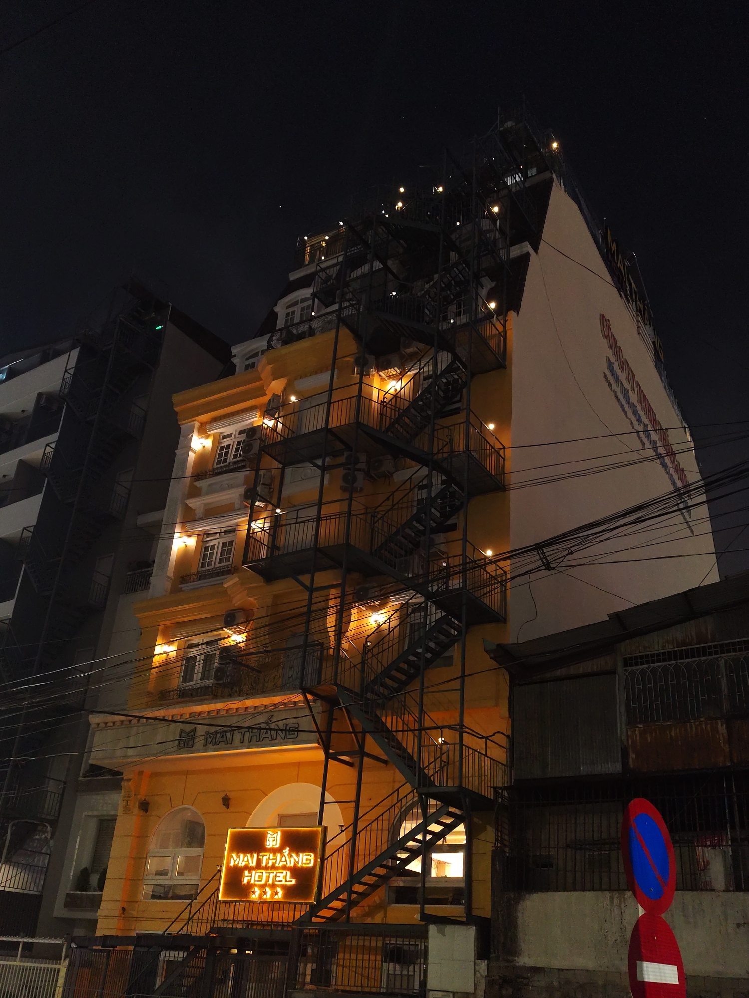 At night in Da Lat, Vietnam, a yellow building stands illuminated. A black metal fire escape climbs its facade. The sign reads "Mai Thang Hotel ***". Another taller building is behind the hotel, also with a black metal fire escape. A stop sign is visible in the foreground. Overhead wires crisscross the scene.