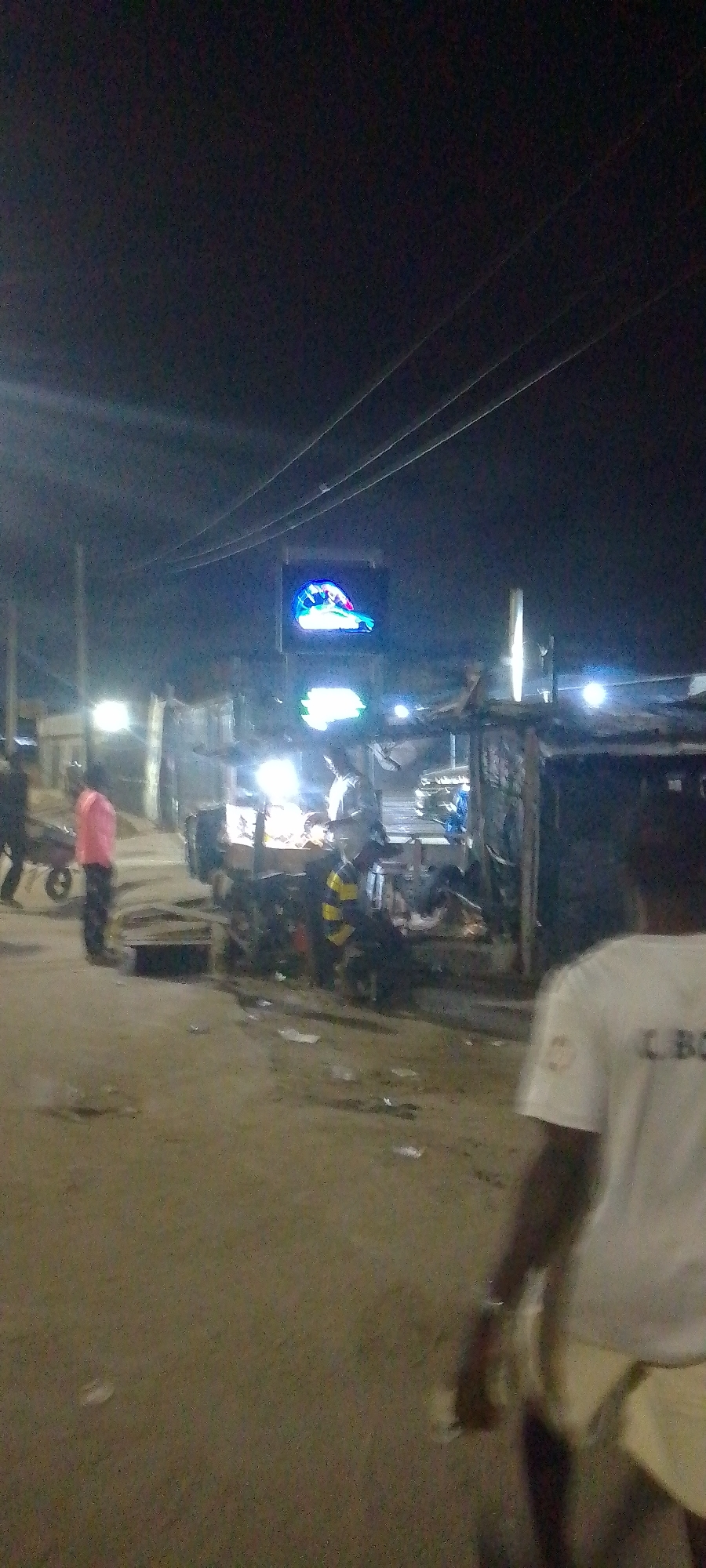 The image displays an unpaved urban street scene at night in Unknown, Unknown. Overhead power lines are visible across the upper portion of the frame. The ground consists of dirt or sand, with scattered debris. Several bright, artificial light sources illuminate the area, primarily originating from structures on the right side. On the right, multiple informal market stalls or structures, constructed from basic materials, line the street. A prominent blue and white illuminated sign, depicting a stylized object resembling a hand or glove, is visible in the mid-ground above these structures.
Approximately five individuals are present. In the immediate foreground on the right, a person with their back to the viewer wears a white t-shirt featuring dark lettering on the back. To the mid-ground left, a person in a pink shirt stands, facing towards the right. Behind them, another individual in dark clothing pushes a two-wheeled cart. Near the market stalls on the mid-ground right, a person in a yellow and black striped shirt is seated, with additional indistinct figures present around the brightly lit stalls.