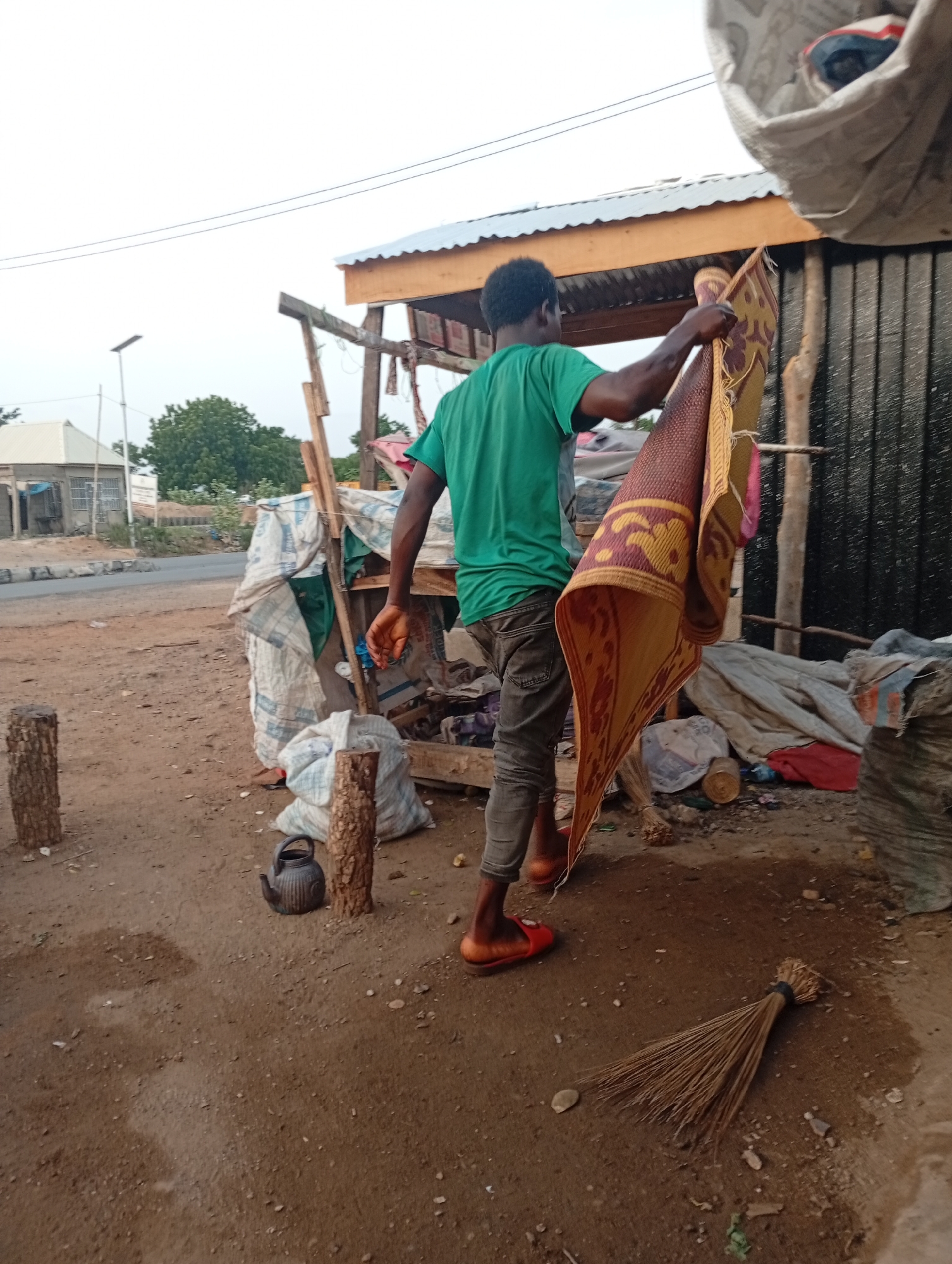 The image shows a person in Adankolo, Nigeria holding up a rectangular mat with brown, yellow, and red geometric patterns. The person is wearing a green t-shirt, gray pants, and orange sandals. The location appears to be a roadside area, with a makeshift structure built with wooden posts and corrugated metal roofing in the background. Various bags and debris are scattered around the ground. A broom lies on the ground in the foreground. There is also a kettle on the ground next to wooden posts. A building is visible in the background across a road.