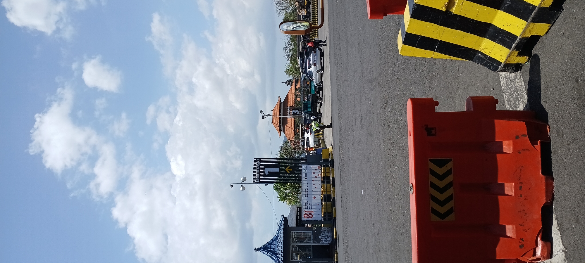 The image captures a view of a ferry terminal in Melaya, Indonesia, on a clear day with scattered clouds. In the foreground, there are two traffic barriers: a bright orange one on the left and a black and yellow striped one on the right. Beyond the barriers, a paved area serves as a loading and unloading zone.
Further into the scene, a blue and grey toll booth or ticket office is visible on the left. To its right, a large sign displaying "Dermaga 1" (Pier 1) with an upward-pointing yellow arrow is prominently featured. Behind the sign, there are several vehicles, including white cars and a pickup truck. People in uniforms, possibly port or security personnel, are seen directing traffic or performing duties.
In the background, a traditional Balinese-style building with an orange tiled roof stands out, possibly administrative offices or a waiting area. The surrounding environment appears to be a coastal area, with hints of distant hills or mountains. The overall impression is of an active transportation hub.